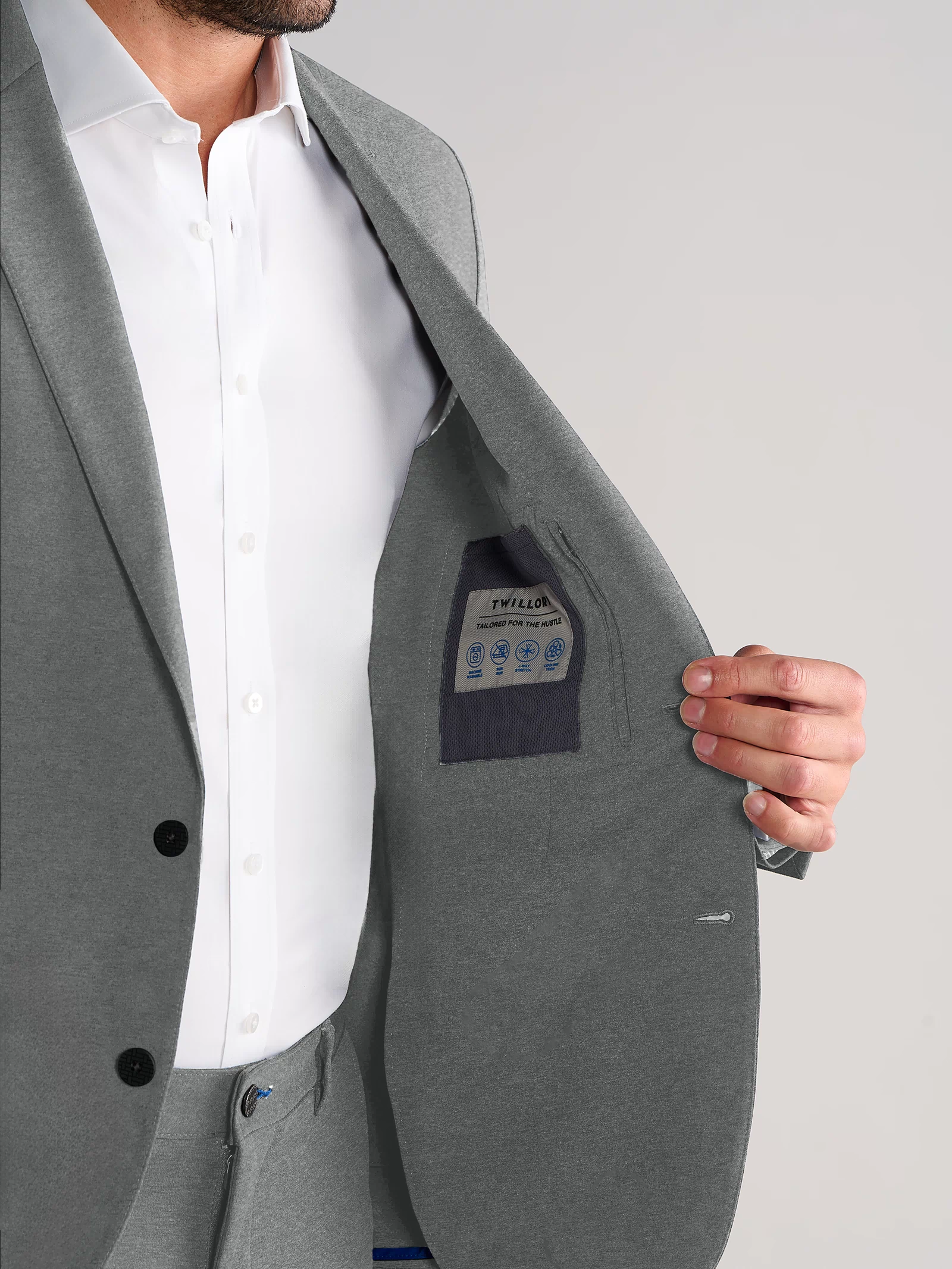 A man in a Twillory Performance Blazer // Heather Light Grey and white shirt holds open his jacket to reveal the inside pocket and label. His face is partially out of frame against a plain light grey background.
