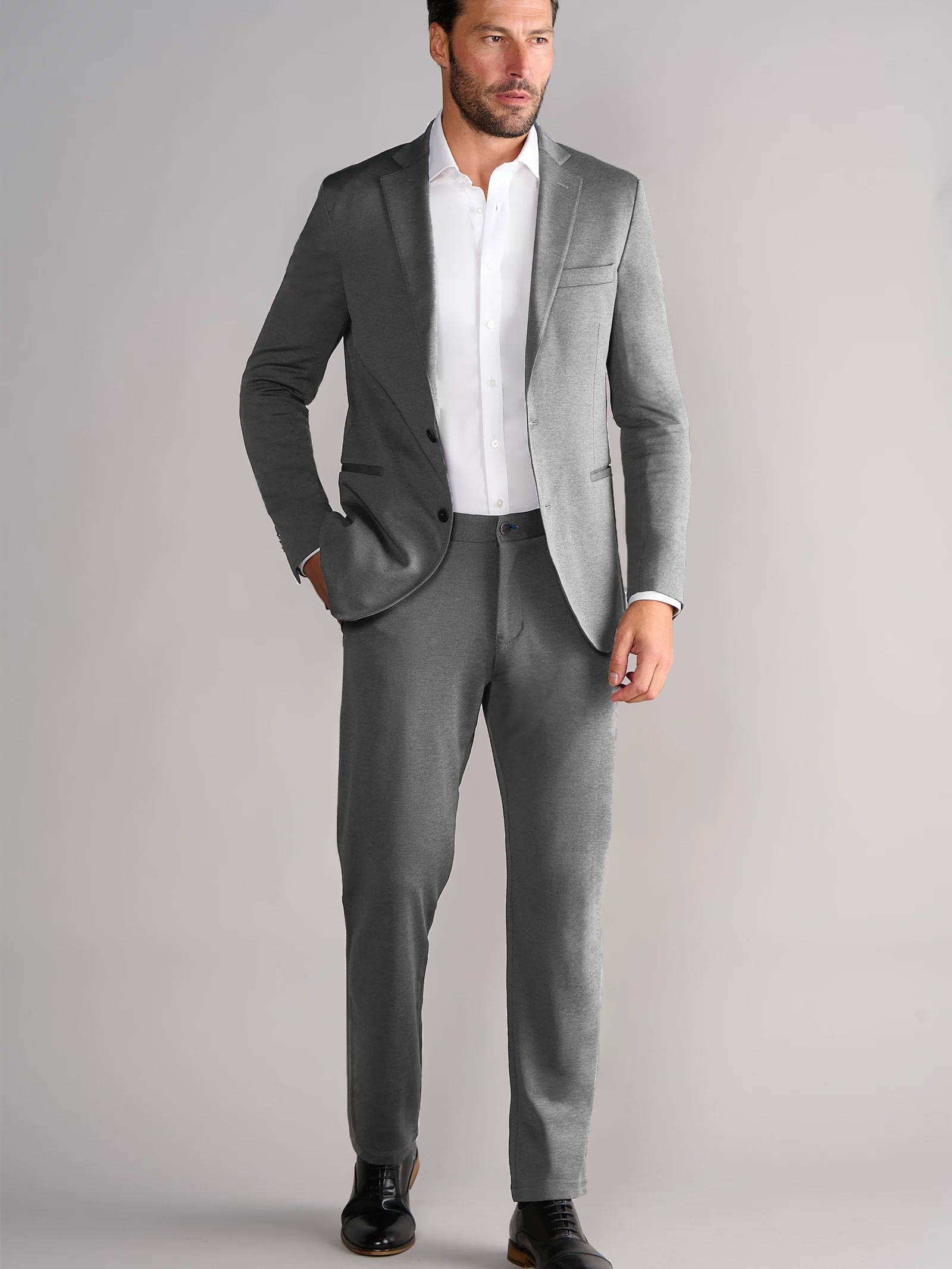 A man with dark hair and a trimmed beard models the Twillory Performance Blazer in Heather Light Grey, paired with a white dress shirt and black shoes. He stands against a light gray backdrop, one hand in his pocket, gazing to the side.