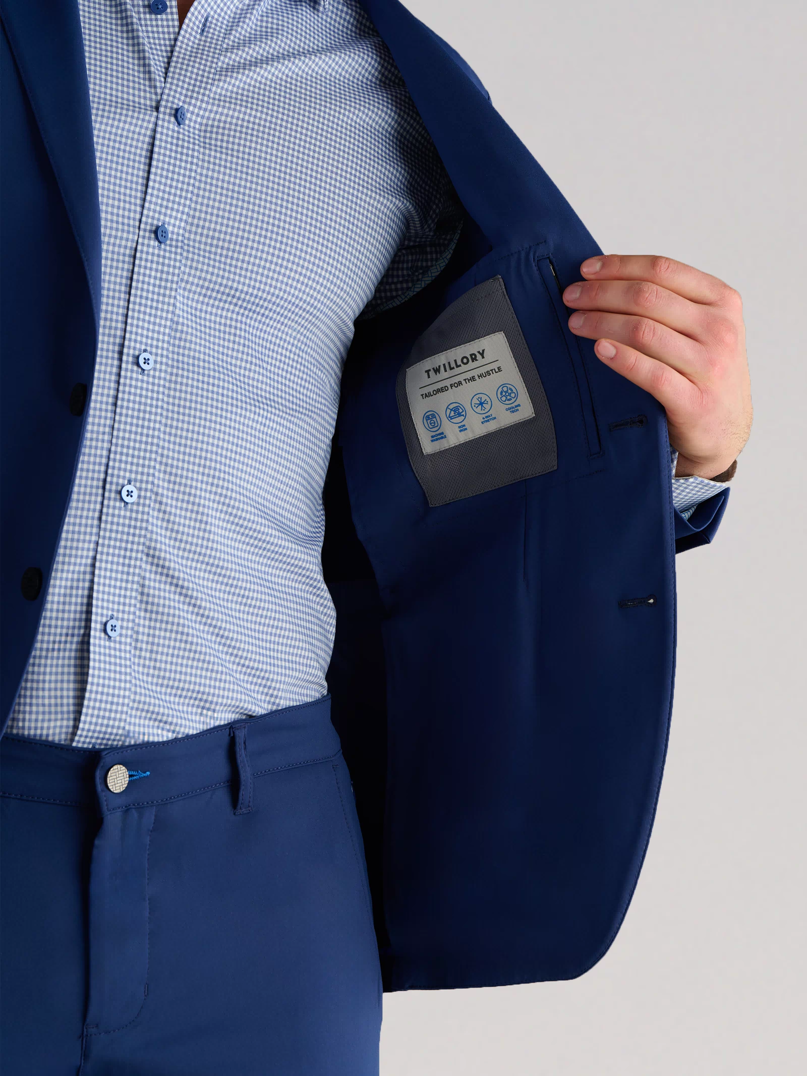 A person wearing the Twillory Performance Blazer in Cobalt Blue and a light blue checkered shirt holds open the jacket to show the inner pocket with a sewn-in label. The background is plain and light-colored.