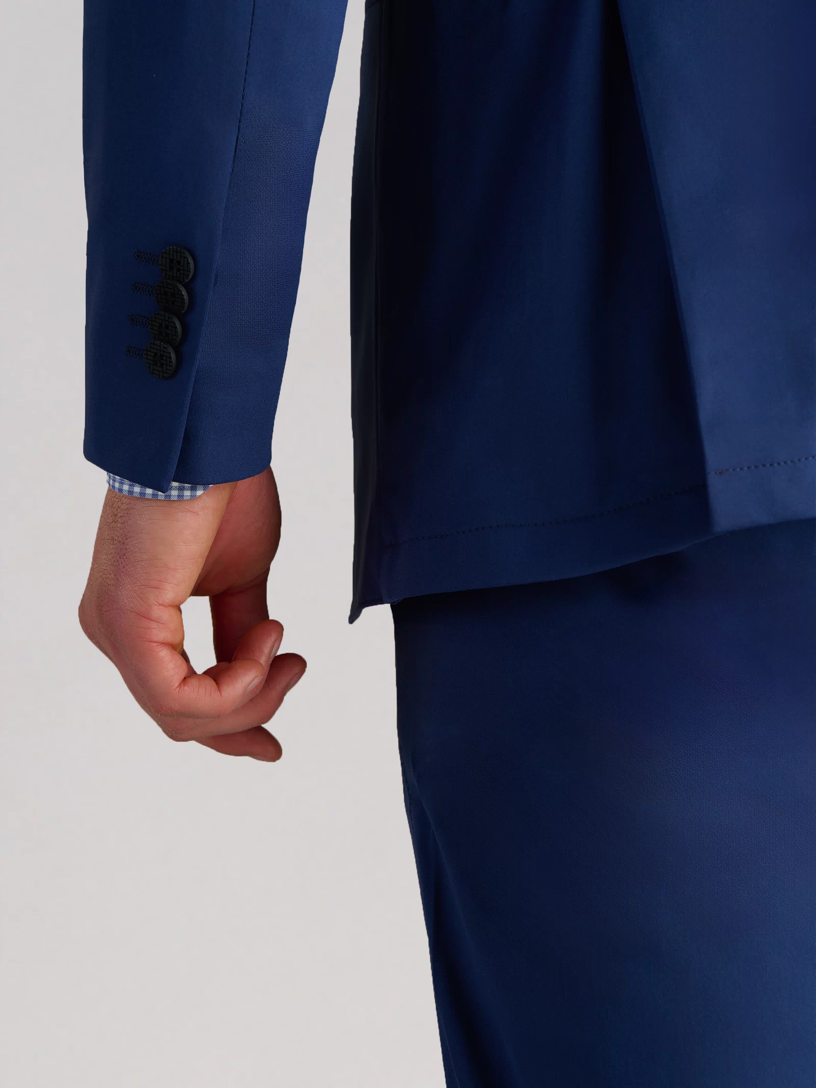 A person wears the Twillory Performance Blazer in Cobalt Blue with matching pants. Their left arm is down, revealing a blue and white check shirt cuff. The background is plain and light-colored.