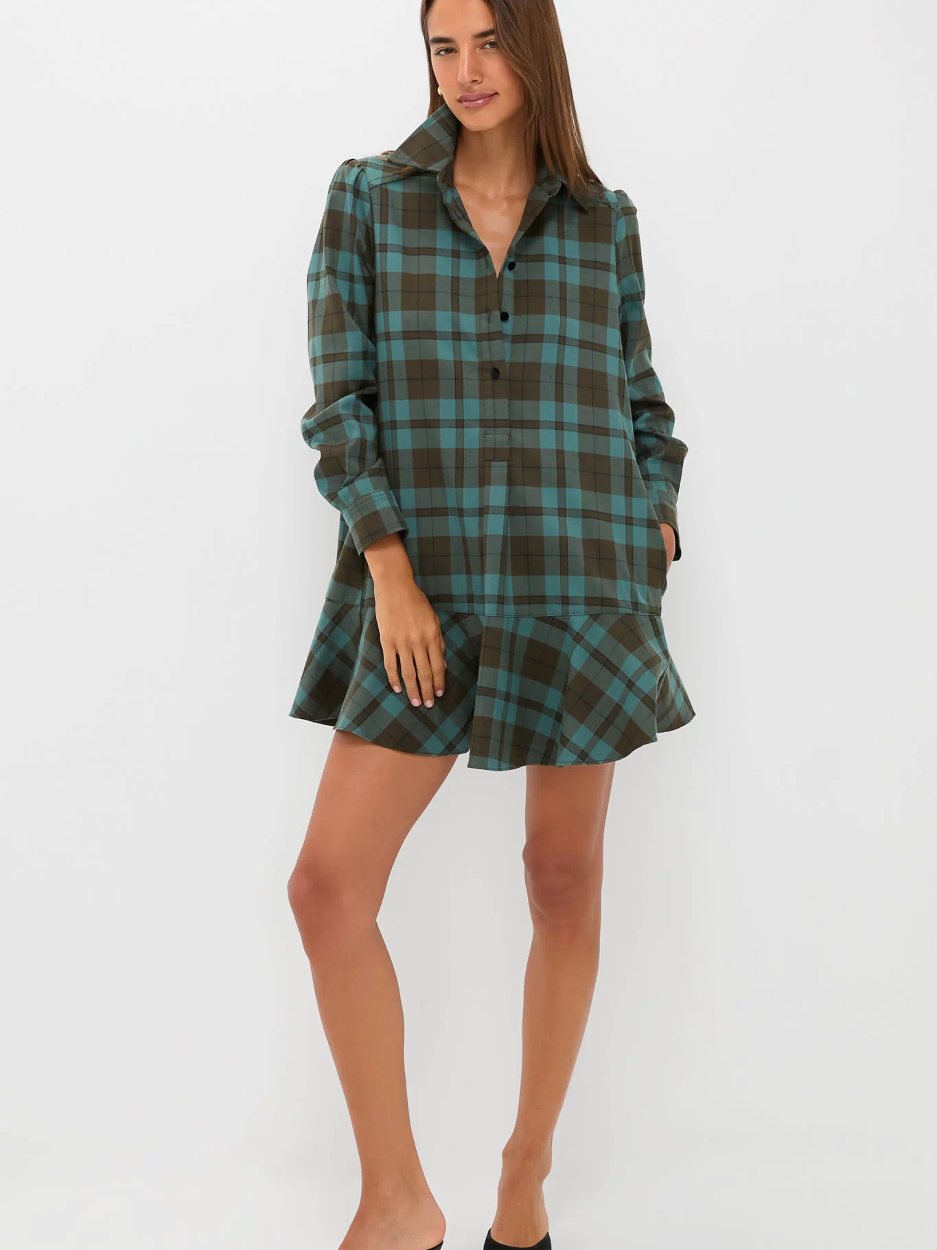 A woman poses against a white background wearing a Tuckernuck brown and teal plaid crepe shirt dress with a flared hem and relaxed fit, styled with black heeled mules.