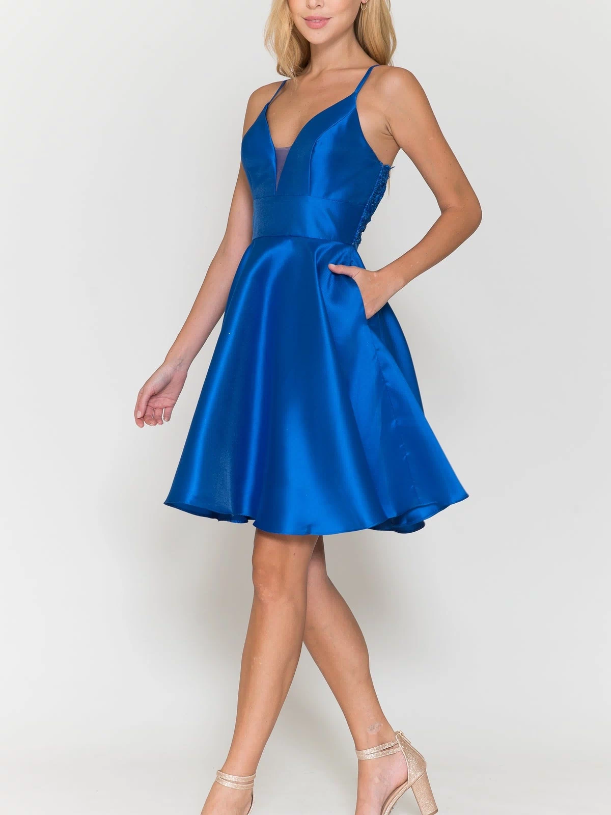 A woman wears the Lace-Back Short A-Line Homecoming Dress 8698, a sleeveless, knee-length royal blue dress with a V-neckline and side pockets, paired with nude high-heeled sandals, standing against a white background.