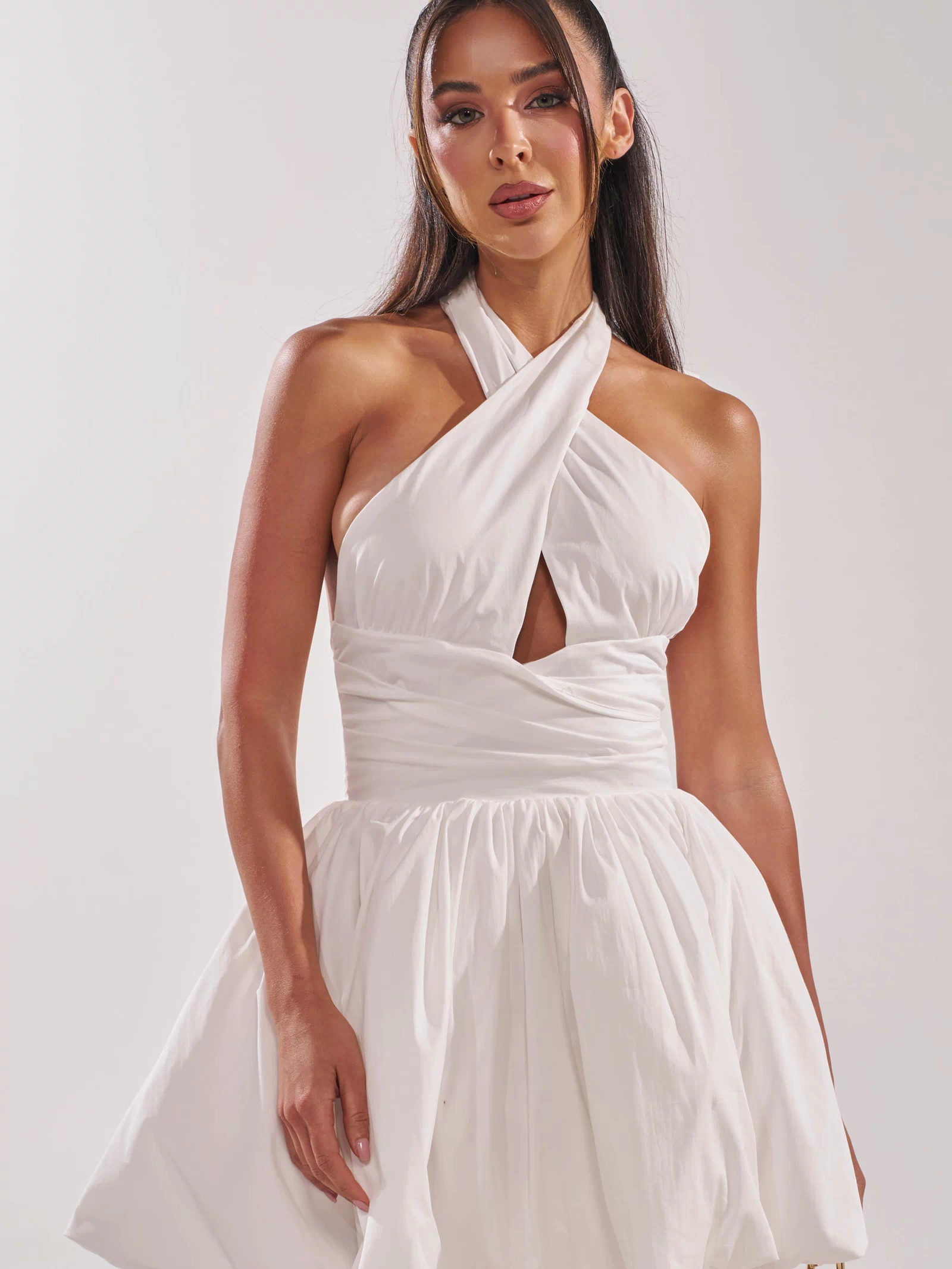 A woman stands against a plain, light background wearing the PRETTY IN POPLIN STRAPPY MULTIFUNCTION MINI DRESS IN WHITE, featuring a sleeveless design, halter neck, bubble hem, keyhole cutout, and short voluminous skirt.