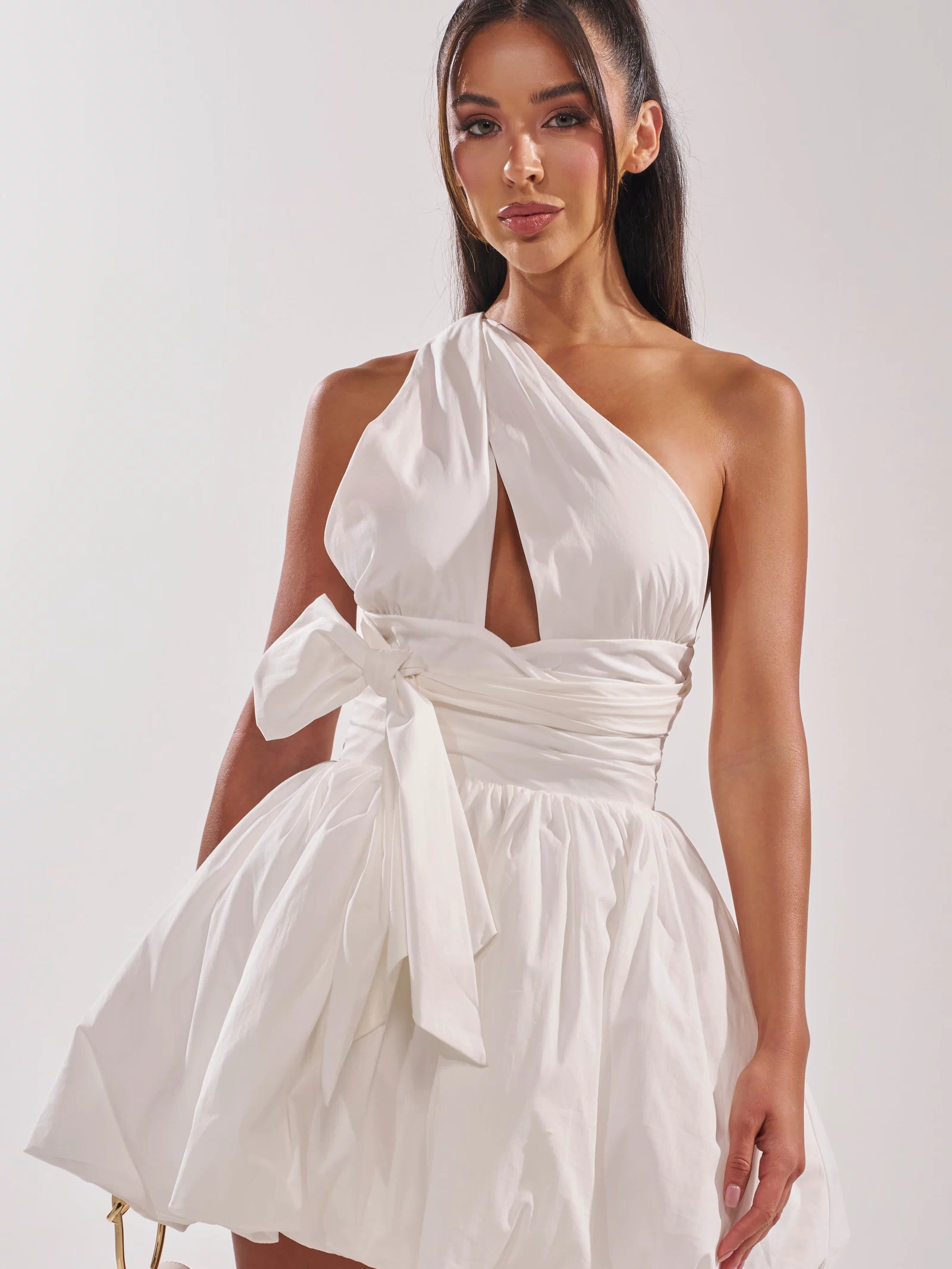 A woman stands against a plain light background, holding a small handbag and wearing the PRETTY IN POPLIN STRAPPY MULTIFUNCTION MINI DRESS IN WHITE featuring a front cutout, waist bow, and flared skirt.