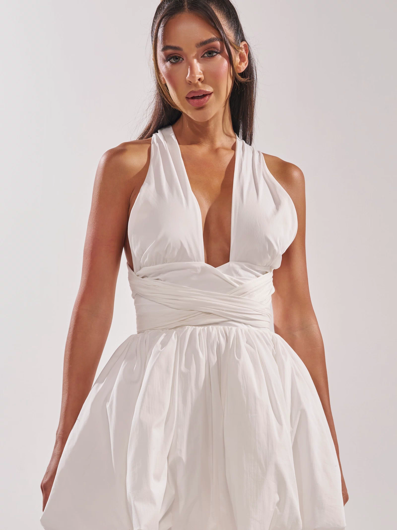 A woman with long dark hair in a half-up style wears the PRETTY IN POPLIN STRAPPY MULTIFUNCTION MINI DRESS IN WHITE, featuring a sleeveless, deep V-neck design, fitted waist, and bubble hem skirt. She stands against a plain light background.