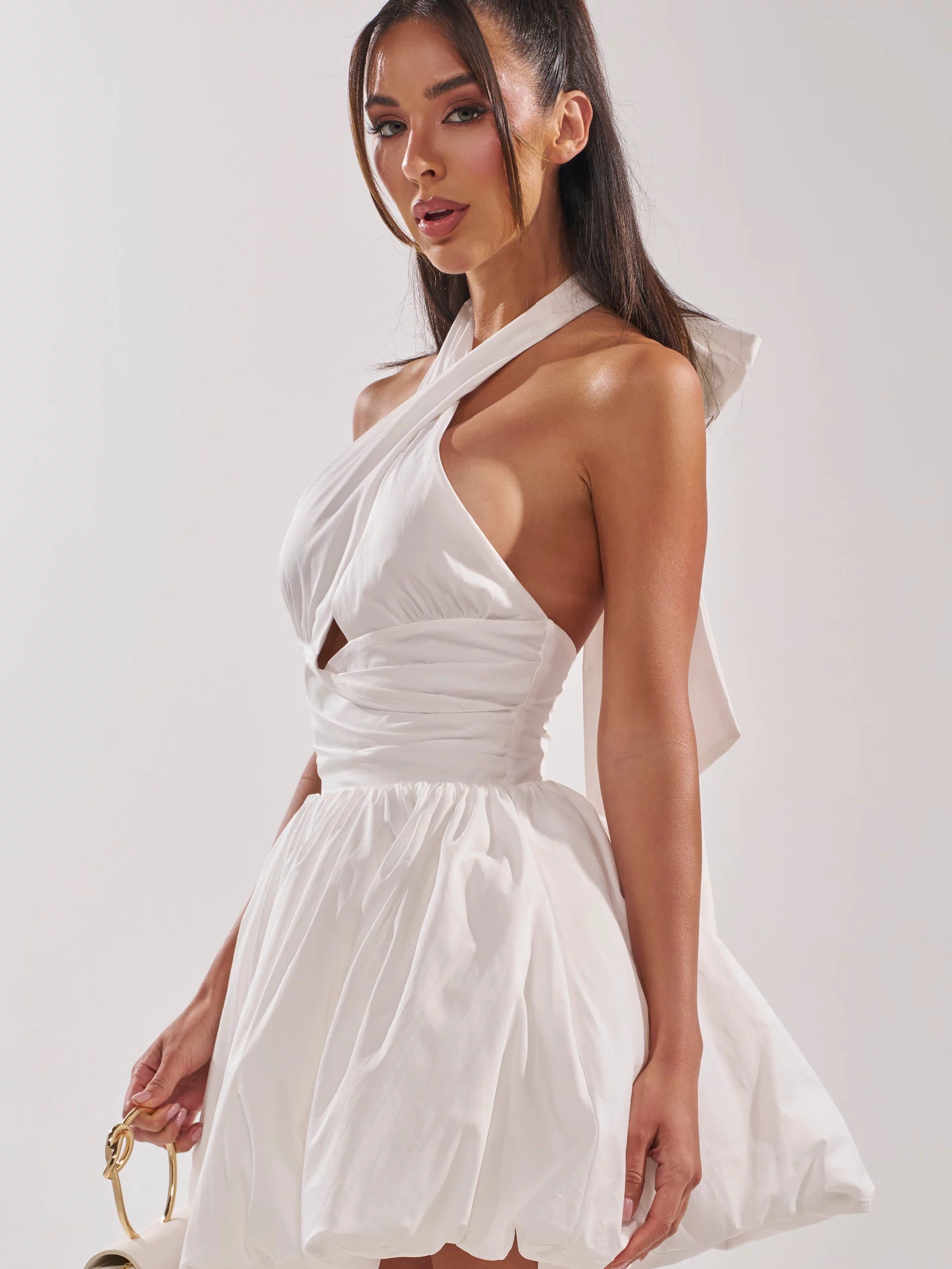 A woman models the PRETTY IN POPLIN STRAPPY MULTIFUNCTION MINI DRESS IN WHITE, featuring a halter neckline and flared skirt. She sports a high ponytail, carries a small white handbag, and stands against a simple light background.