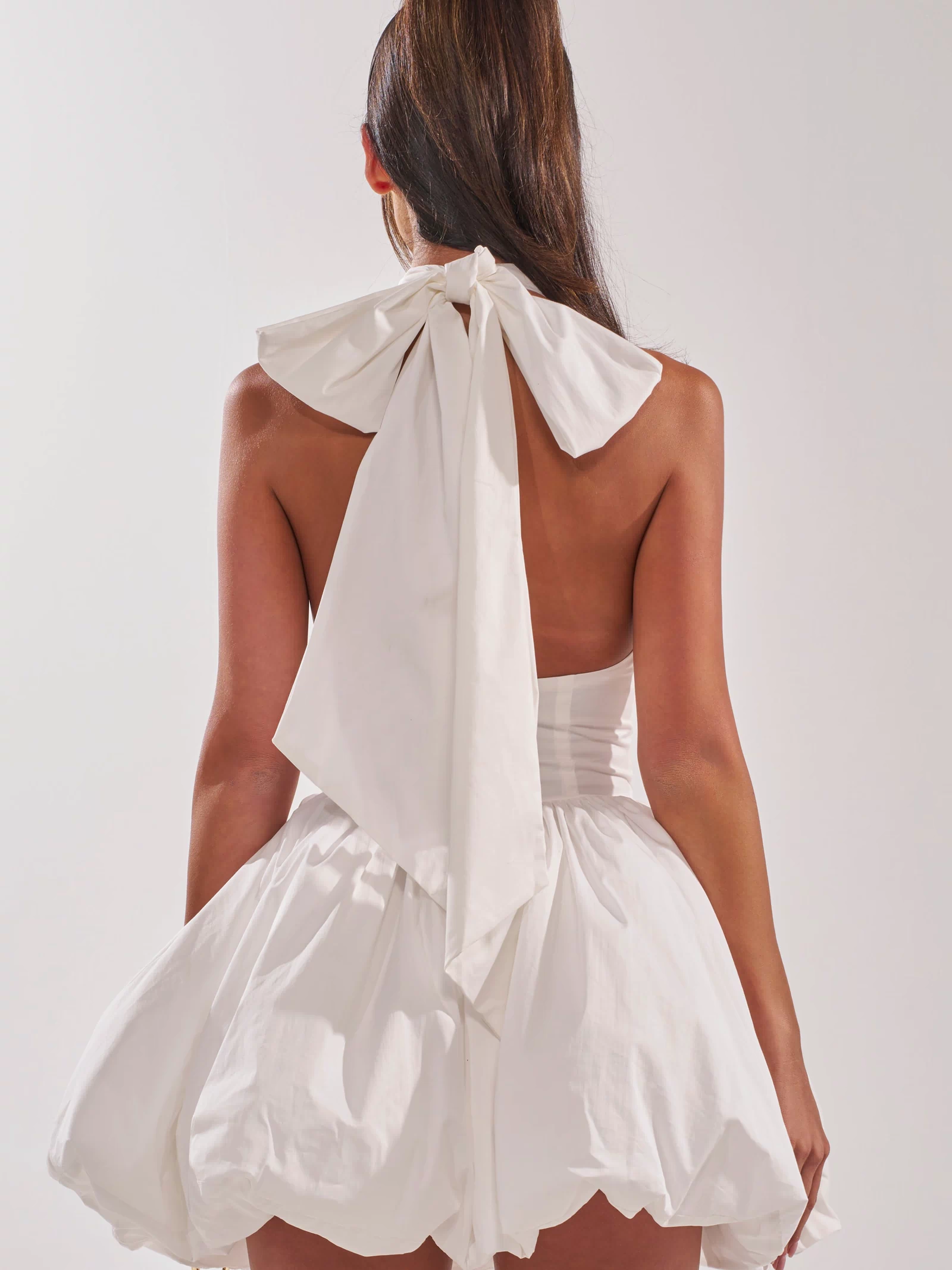 A woman with long brown hair wears the PRETTY IN POPLIN STRAPPY MULTIFUNCTION MINI DRESS IN WHITE, featuring a backless design, large halter neck bow, and voluminous bubble hem skirt, standing against a plain light background.