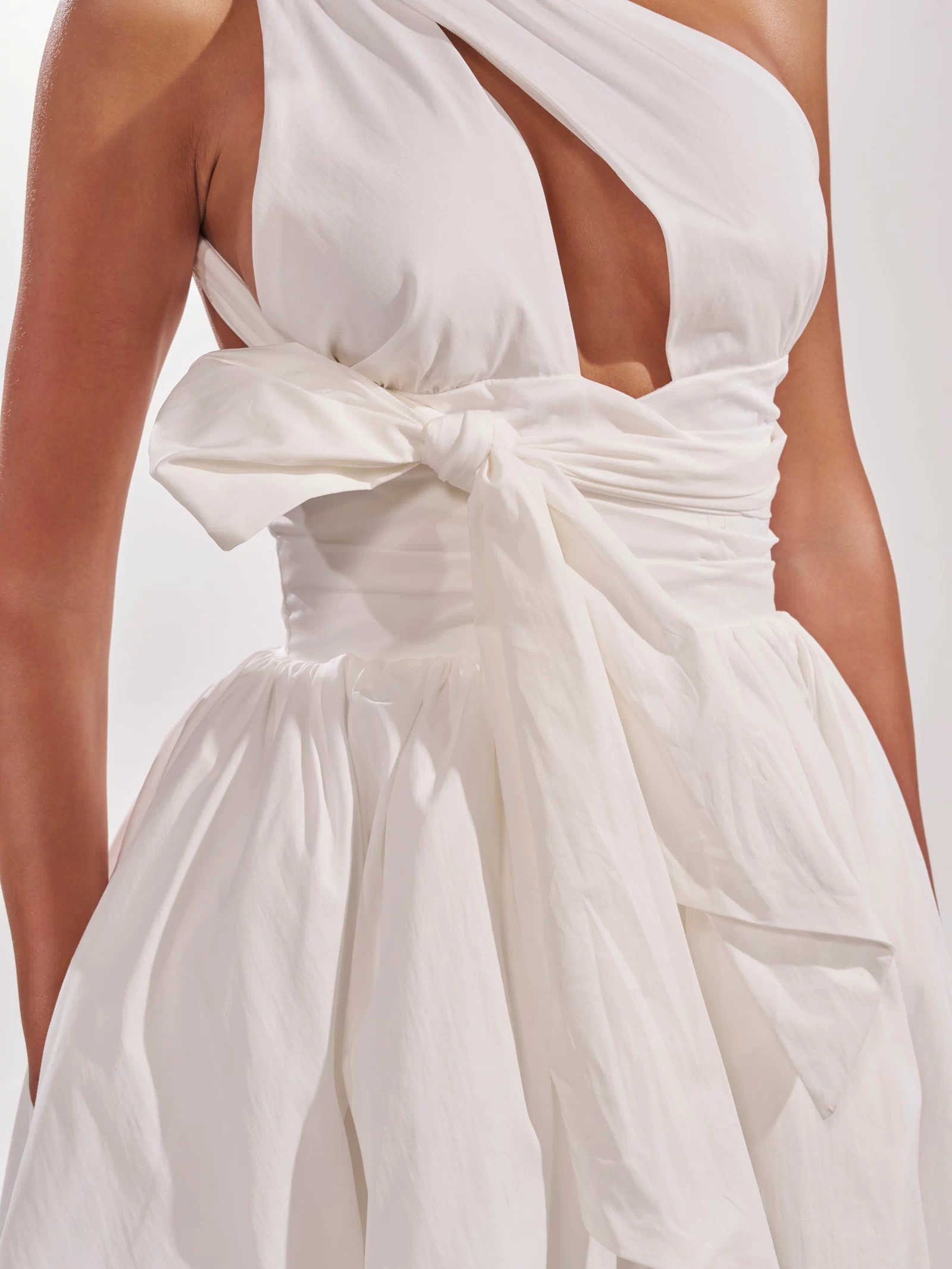 A close-up of a person wearing the PRETTY IN POPLIN STRAPPY MULTIFUNCTION MINI DRESS IN WHITE, featuring a chic one-shoulder cut, front cutout, wide sash bow at the waist, and a softly gathered skirt with a subtle bubble hem.