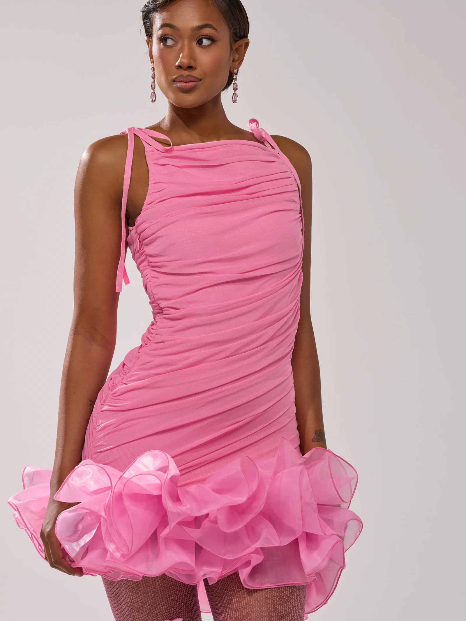 A woman in the PRINCESS PEPLUM MINI DRESS IN PINK—a fitted, ruched mesh mini dress with a ruffled hem—pairs it with pink earrings and fishnet tights, standing confidently against a plain background.