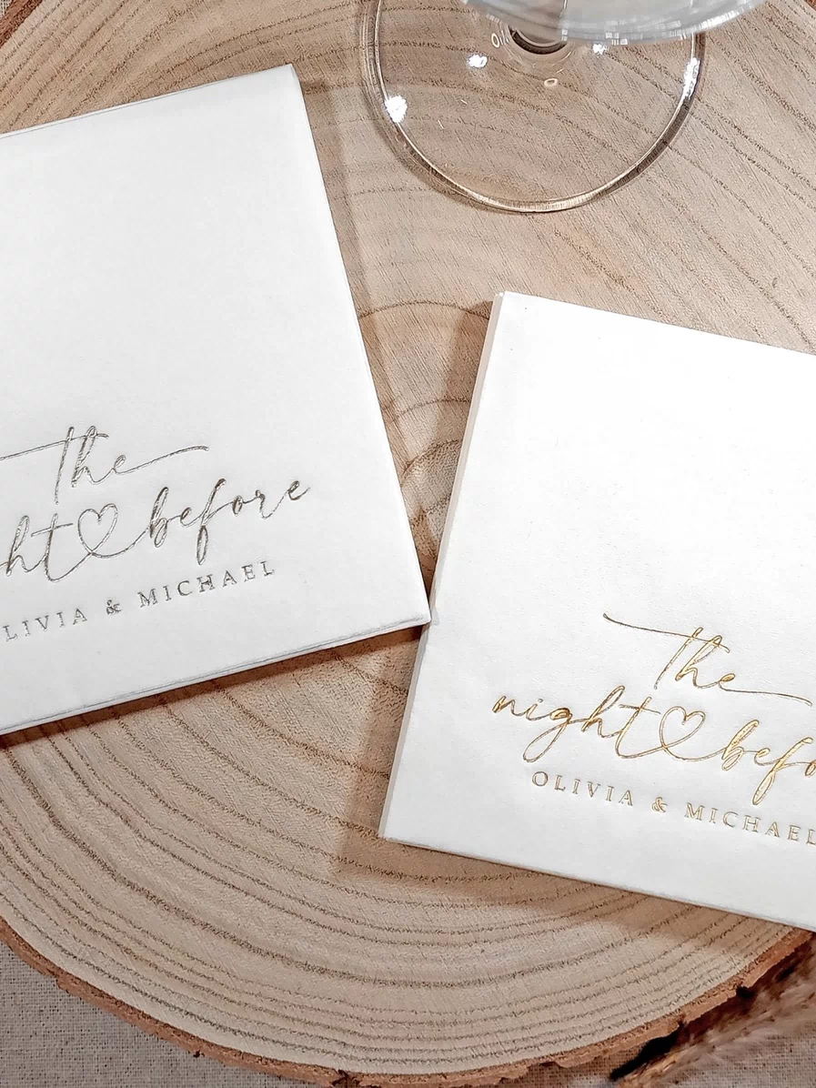 wo stacks of white napkins—one with gold foil and one with silver foil—both featuring “the night before” script with a heart and names.