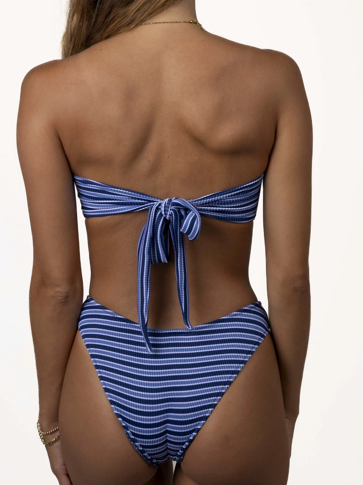 A person wearing the Honey Swim Santorini Ribbed Bandeau in blue multi-stripe is shown from the back. Their long hair drapes over one shoulder, and bracelets adorn their left wrist.