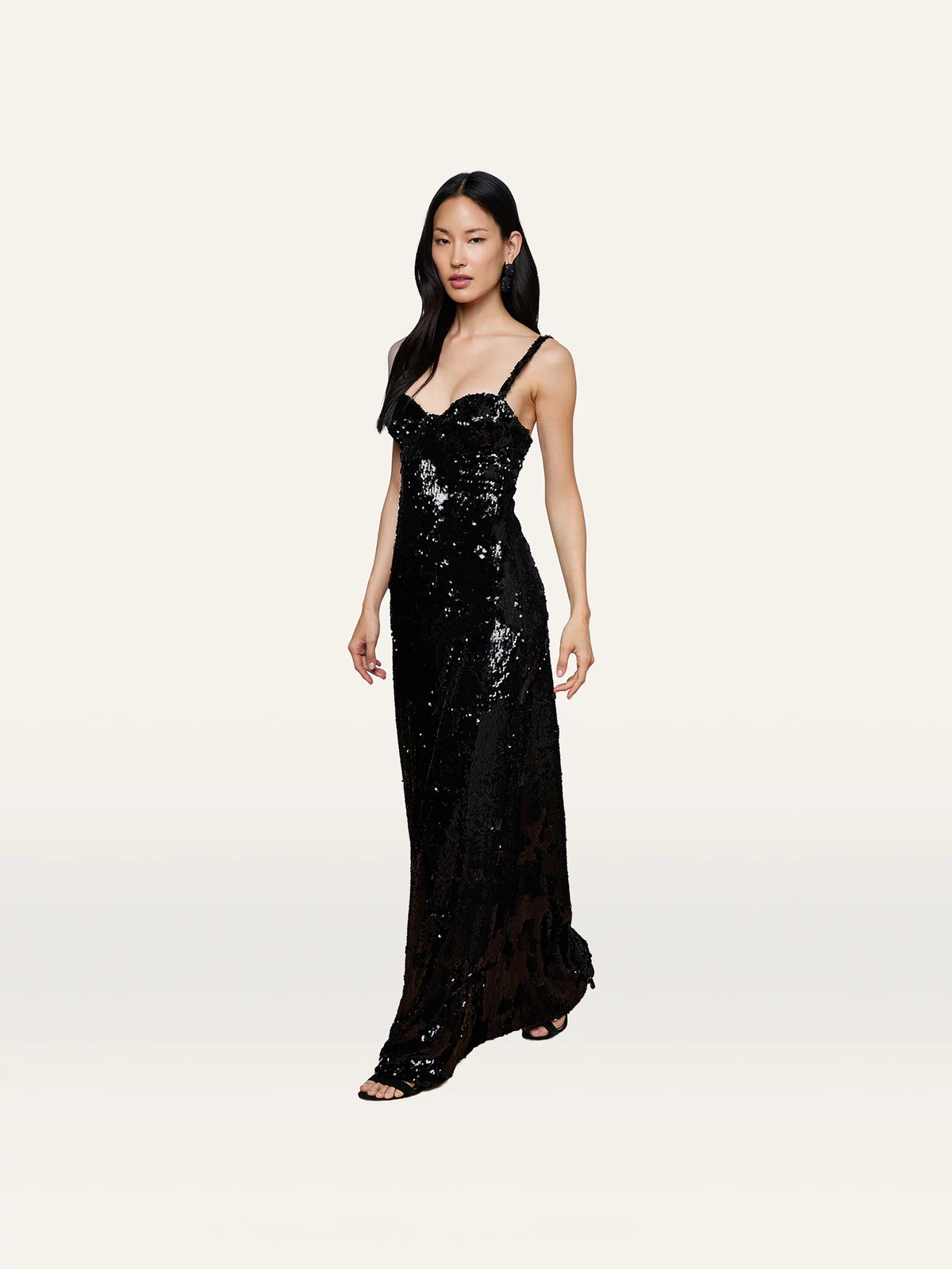 A woman wearing the Sayla Sequin Gown—a long, black sequin dress with thin straps that exudes vintage glamour—stands against a plain light background.