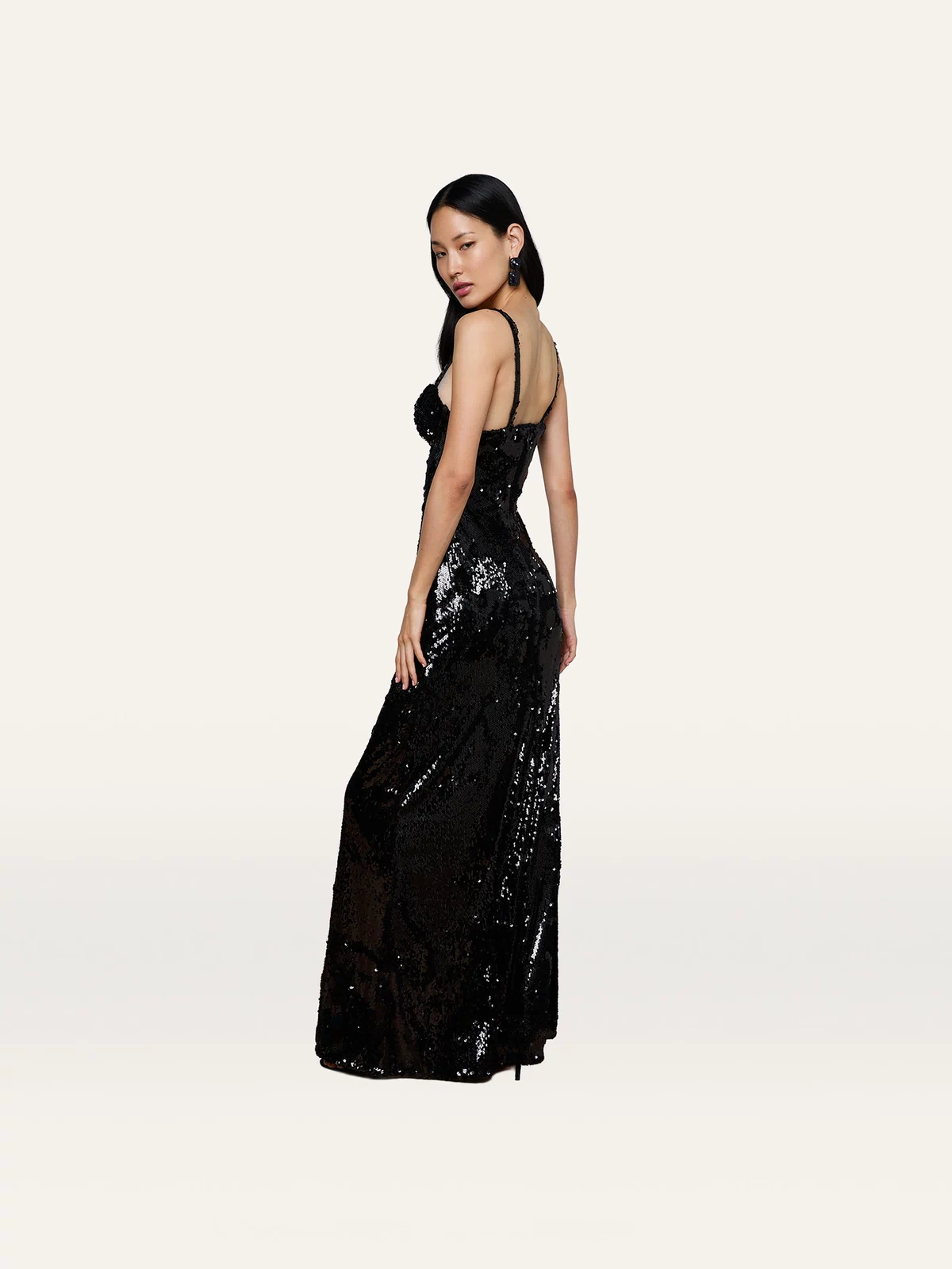 Draped in the Sayla Sequin Gown—a long, sleeveless black sequin dress—a woman stands against a plain light background, channeling vintage glamour as she gazes over her shoulder with one hand resting by her side.
