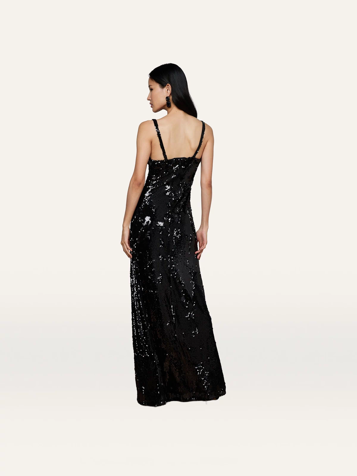 A woman stands facing away against a plain light background, showcasing the back of the Sayla Sequin Gown—a long, sleeveless black sequin dress that exudes vintage glamour.