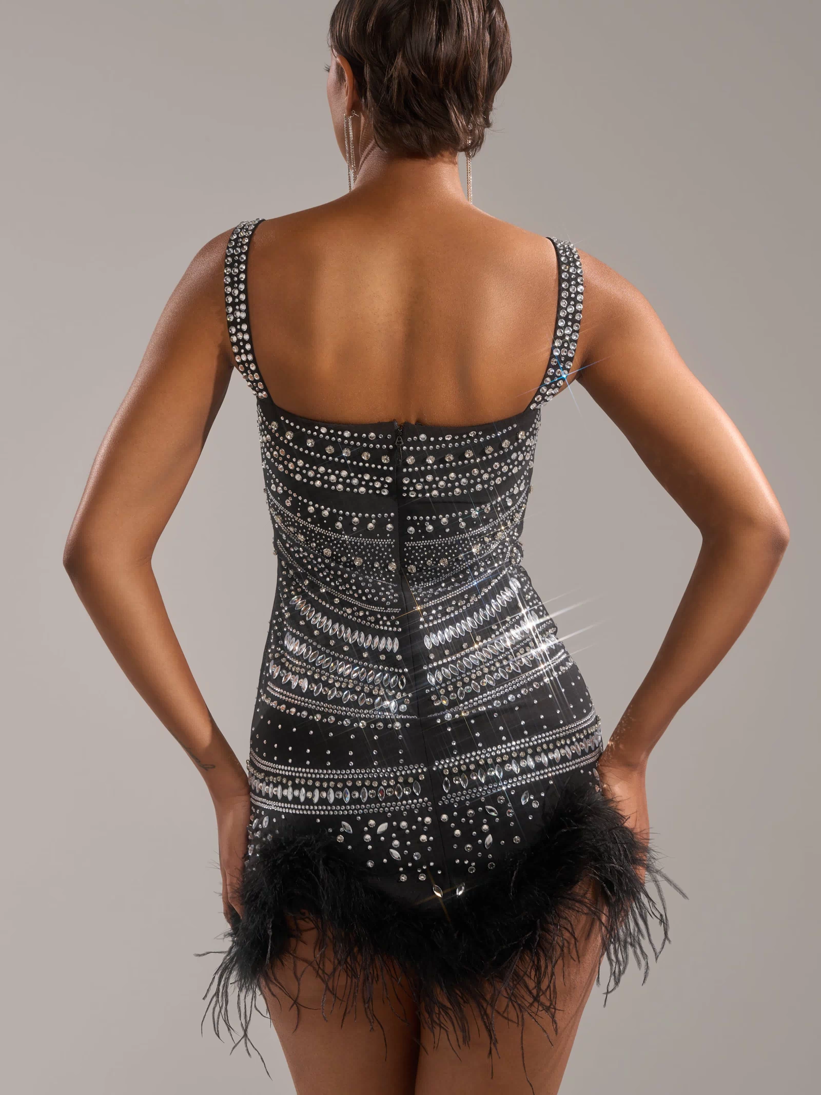 A person faces away wearing the SHOWSTOPPER RHINESTONE MINI DRESS, a black bodycon mini with feather trim and sparkling silver sequins and beads, set against a plain neutral background.