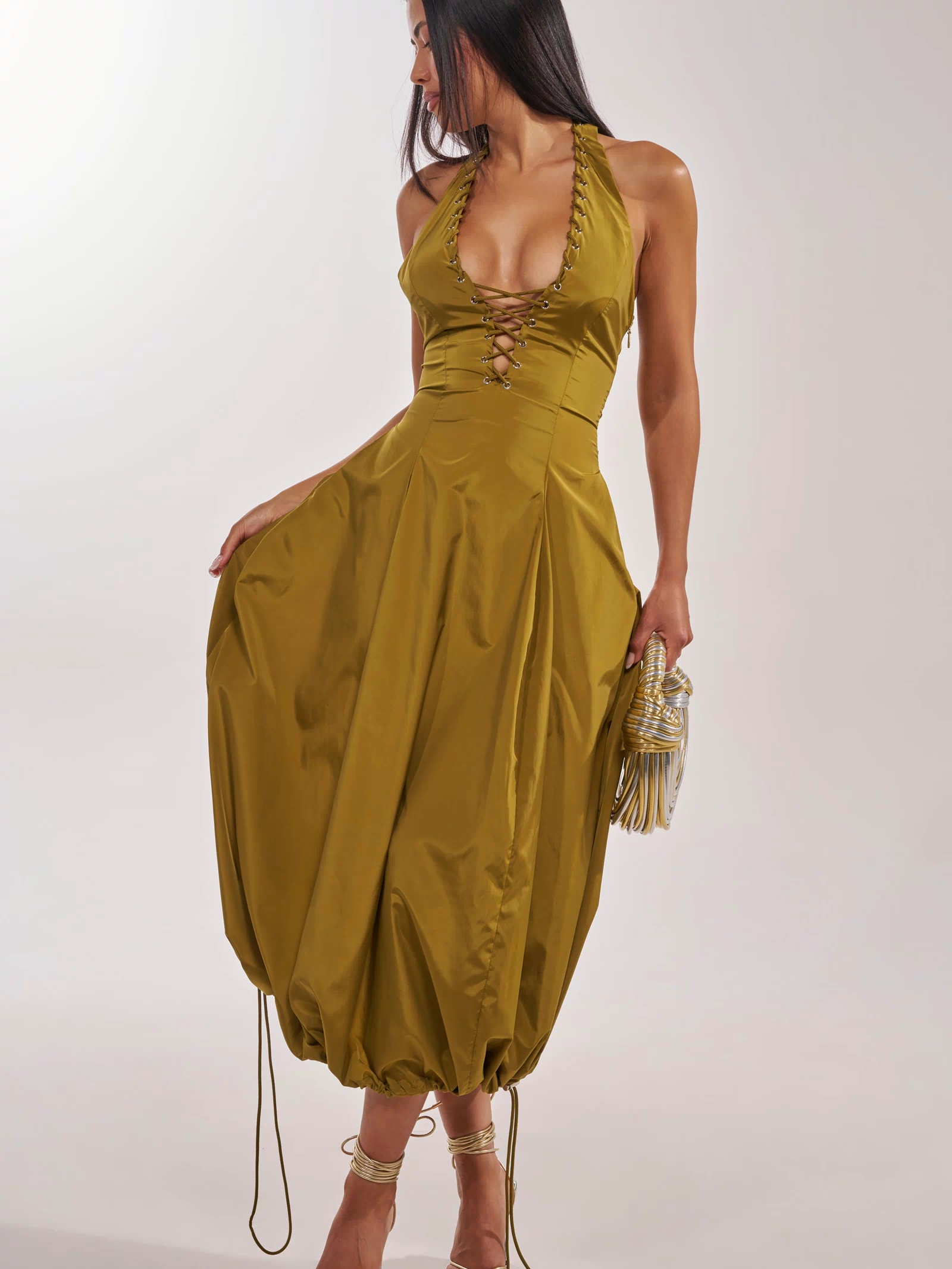 A woman poses in the SOMETHING LIGHT NYLON MAXI DRESS, featuring an olive hue, lace-up front detail, and a voluminous gathered skirt. She accessorizes with a gold clutch and strappy heels.
