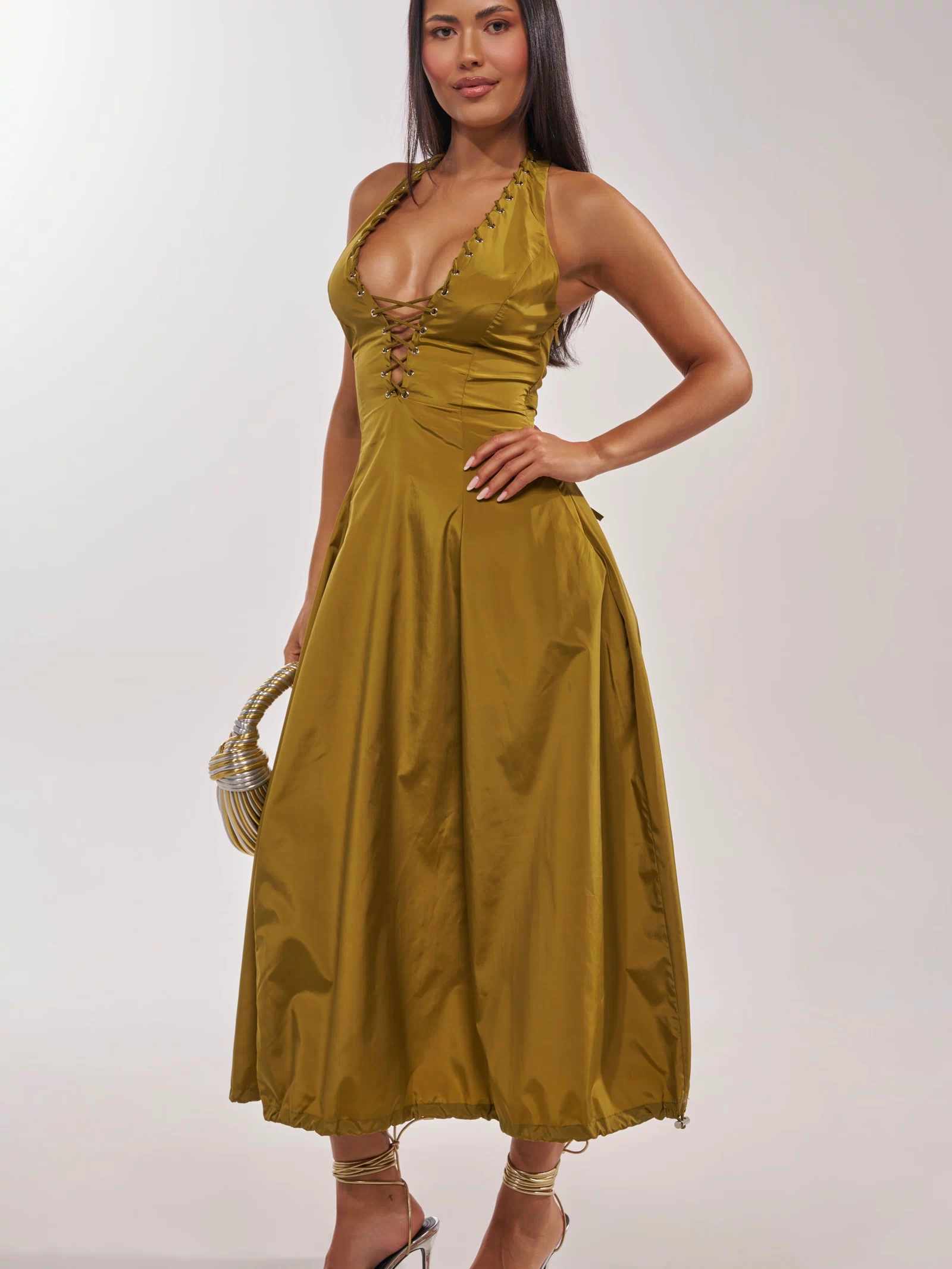 A woman stands confidently in the SOMETHING LIGHT NYLON MAXI DRESS—a sleeveless halter-neck maxi with a gold lace-up detail and mid-length skirt. She completes her look with a small handbag and strappy high-heeled sandals.