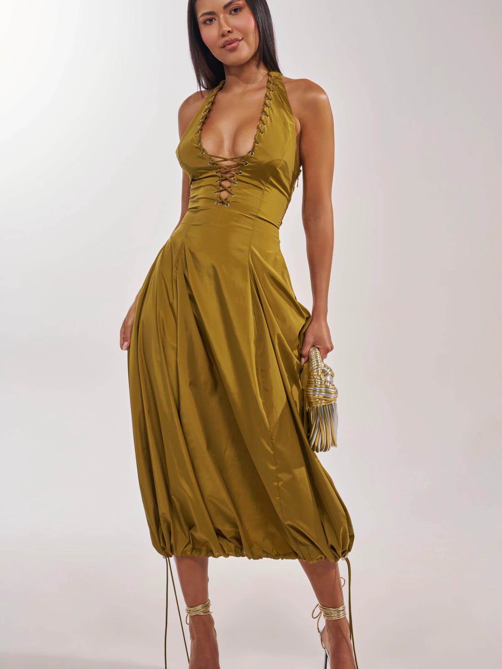 A woman models the SOMETHING LIGHT NYLON MAXI DRESS in shiny olive with a deep neckline and lace-up front, holding a small ruched handbag. She pairs it with strappy high-heeled sandals and wears her straight, dark hair down.