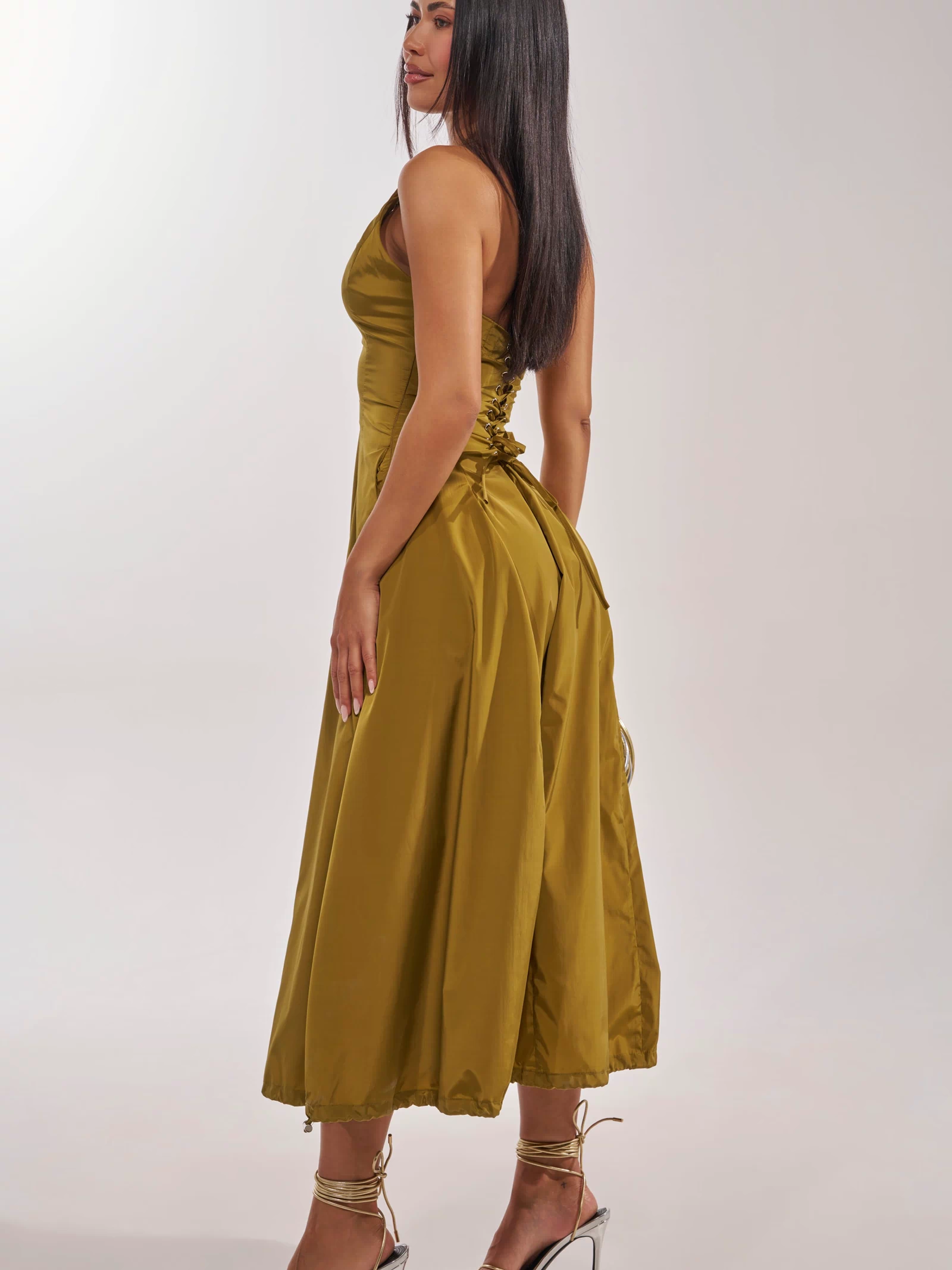 A woman stands sideways in the SOMETHING LIGHT NYLON MAXI DRESS, an olive green, shiny utilitarian piece with a fitted bodice and flared skirt, styled with strappy high-heeled sandals against a neutral background.