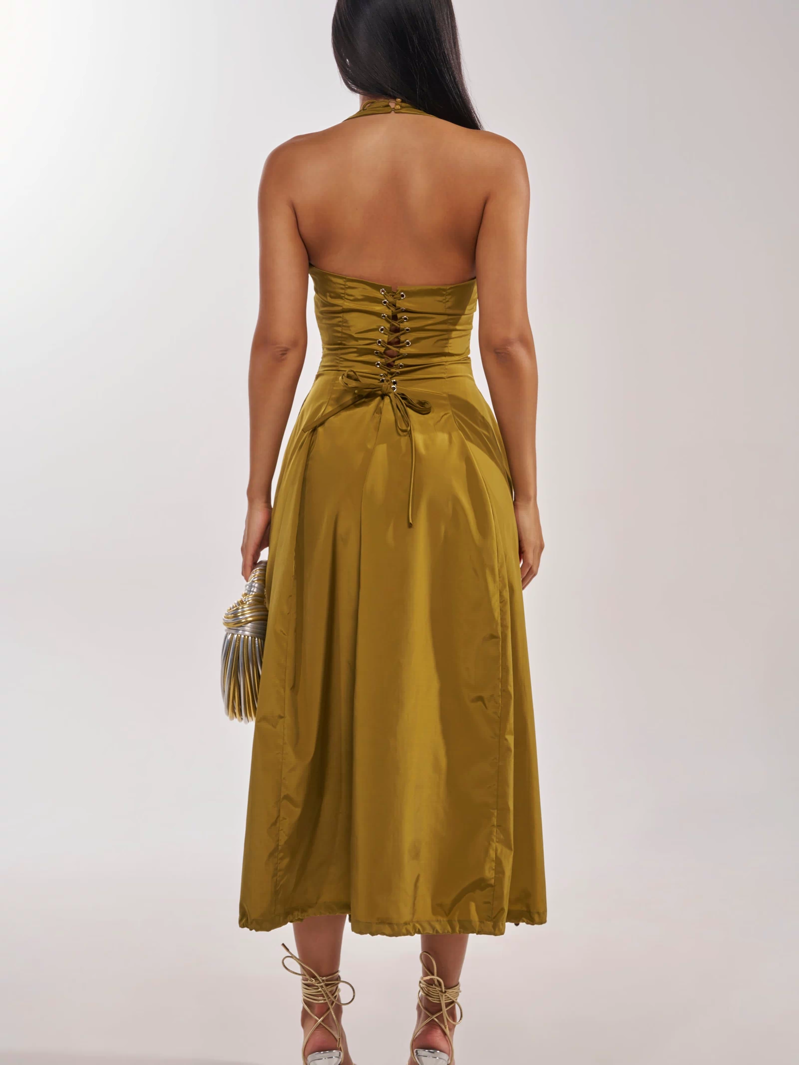 A woman stands with her back to the camera, wearing the SOMETHING LIGHT NYLON MAXI DRESS in shiny olive, paired with a matching fringed bag and strappy high heels.