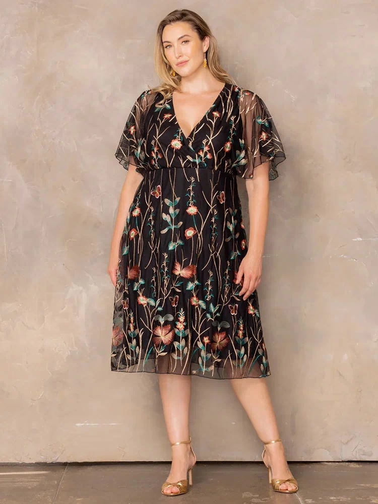 Sonnet Embroidered Midi Cocktail Dress in Onyx Meadow Print  Model wearing black embroidered mesh midi dress with V-neck and flutter sleeves, posing against beige wall in gold heels.