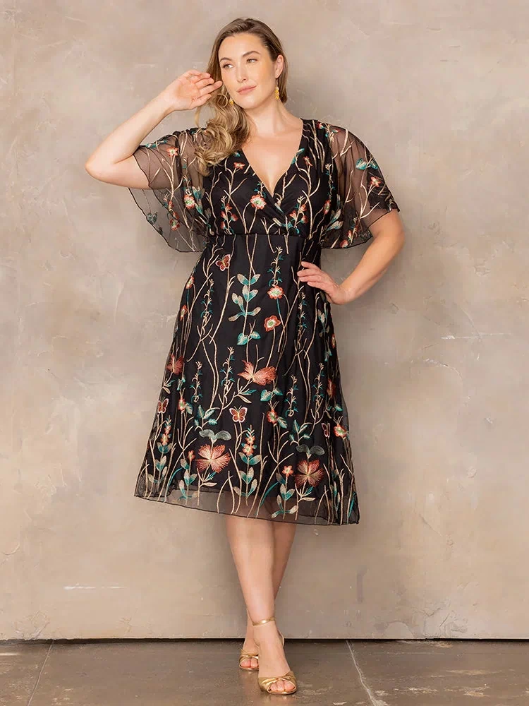 Sonnet Embroidered Midi Cocktail Dress in Onyx Meadow Print  Plus size model wearing black embroidered mesh midi dress with V-neck and sheer flutter sleeves, styled with gold heels for a dressy occasion.