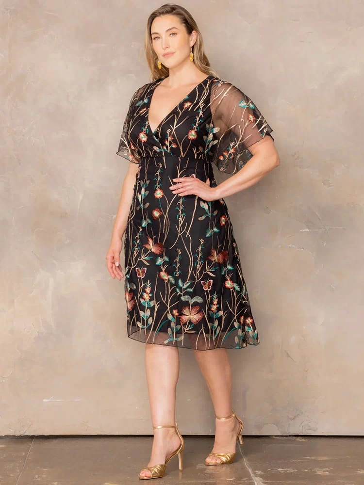Sonnet Embroidered Midi Cocktail Dress in Onyx Meadow Print  Plus size model wearing black embroidered mesh midi dress with V-neck and sheer flutter sleeves, posing in gold heels against a neutral backdrop.