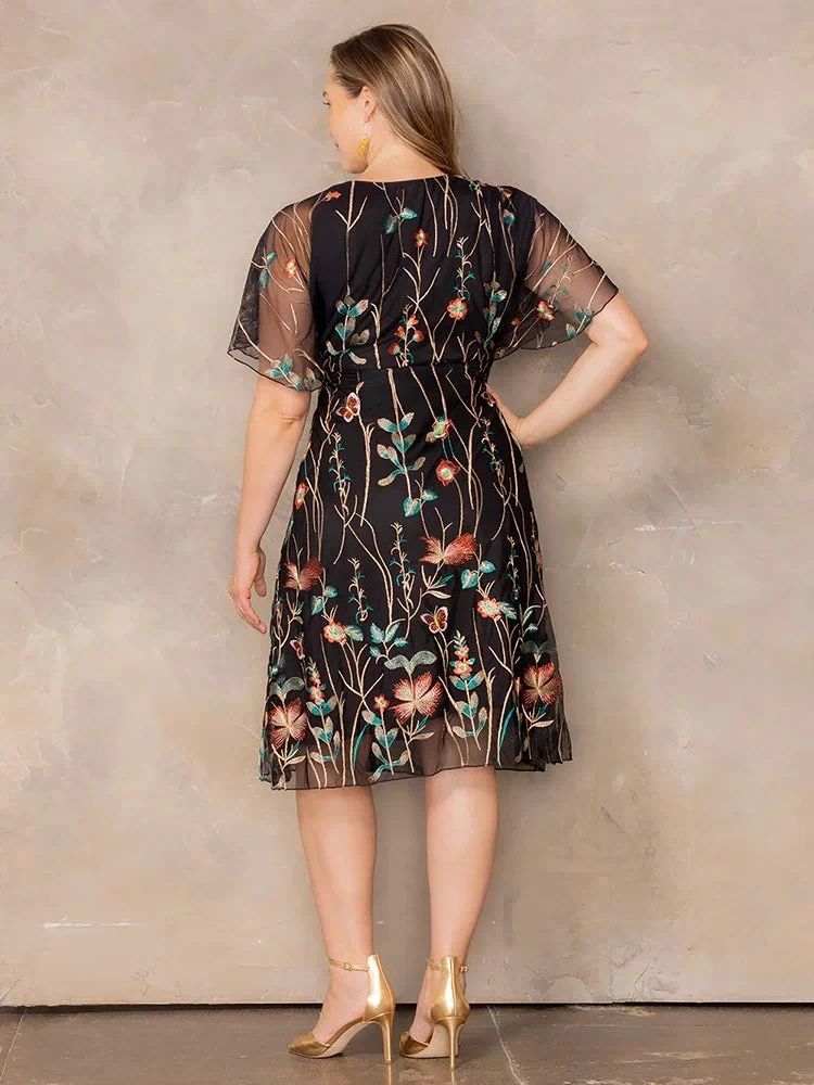 Sonnet Embroidered Midi Cocktail Dress in Onyx Meadow Print  Back view of plus size model wearing black embroidered mesh midi dress with sheer flutter sleeves, standing in gold heels against a neutral backdrop.