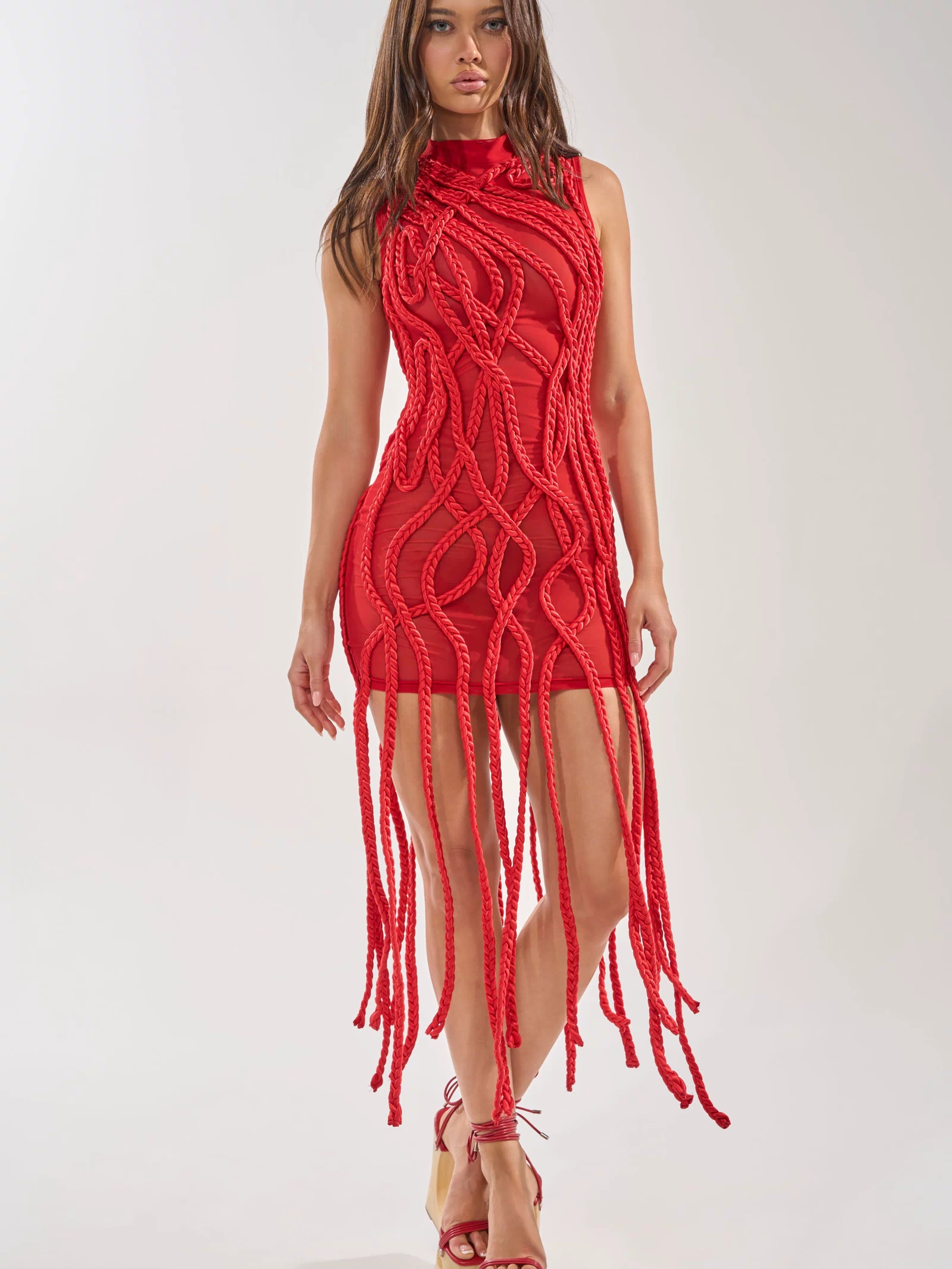 A woman wears the STUCK ON YOU BRAIDED MINI DRESS IN RED, featuring rope-like braided details and a mini length, paired with matching red platform sandals as she stands confidently against a plain white background.