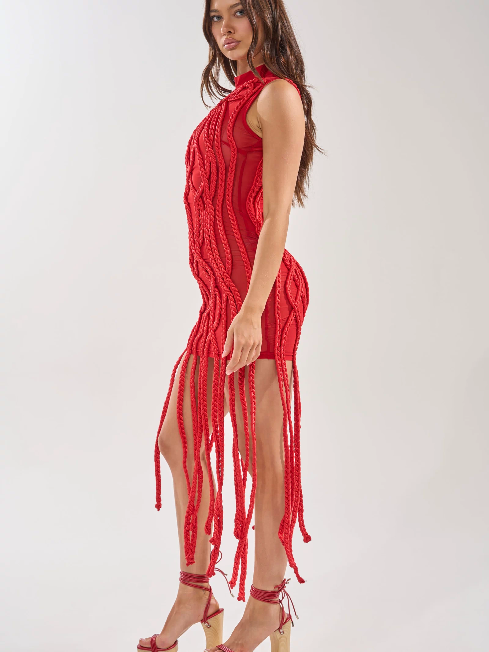 A woman models the STUCK ON YOU BRAIDED MINI DRESS IN RED, featuring bold braided details and long fringe, styled with matching red platform heels. She stands sideways against a light background, looking toward the camera.