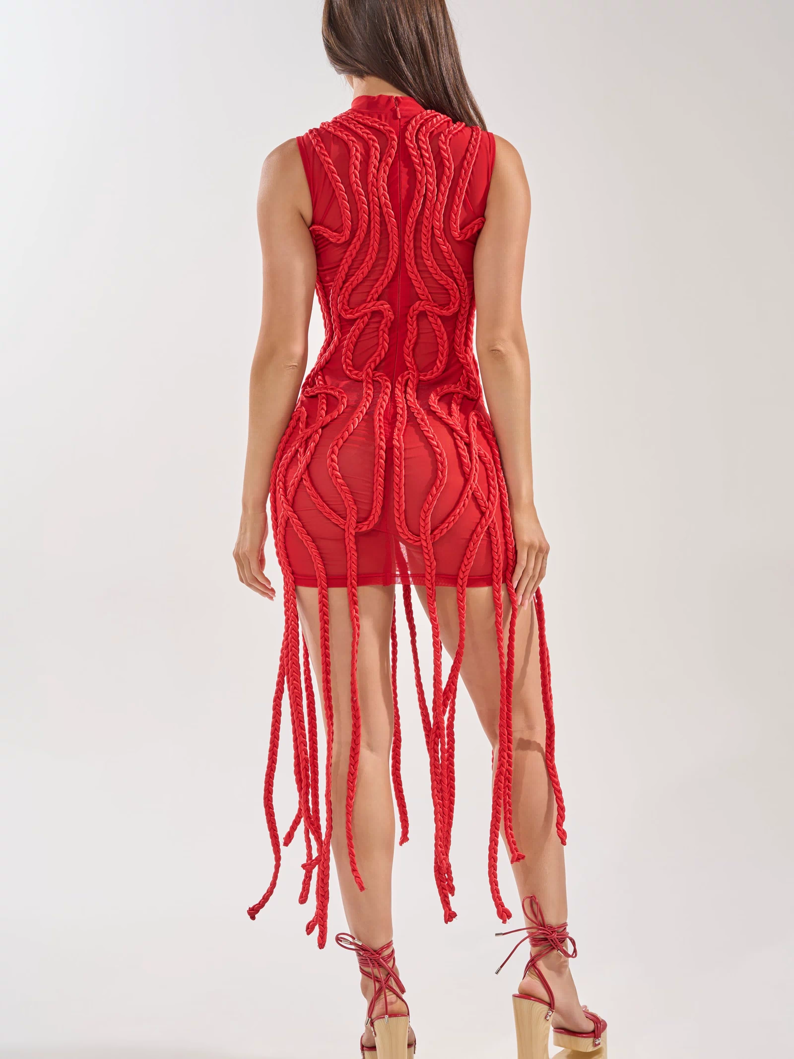 A woman with long brown hair wears the STUCK ON YOU BRAIDED MINI DRESS IN RED, a sleeveless, form-fitting mini dress featuring thick braided details down the back. She pairs it with red lace-up wedge sandals against a plain light background.