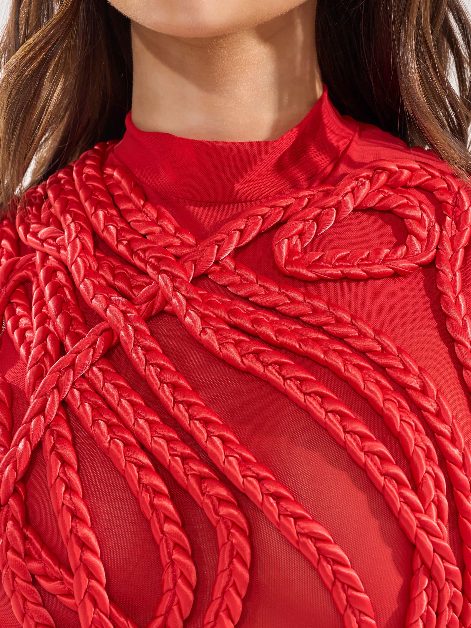 A woman models the STUCK ON YOU BRAIDED MINI DRESS IN RED, featuring sheer fabric and bold, thick braided red ropes in striking patterns across her chest and shoulders. Her lower face and torso are shown, emphasizing the dress’s unique braided details.
