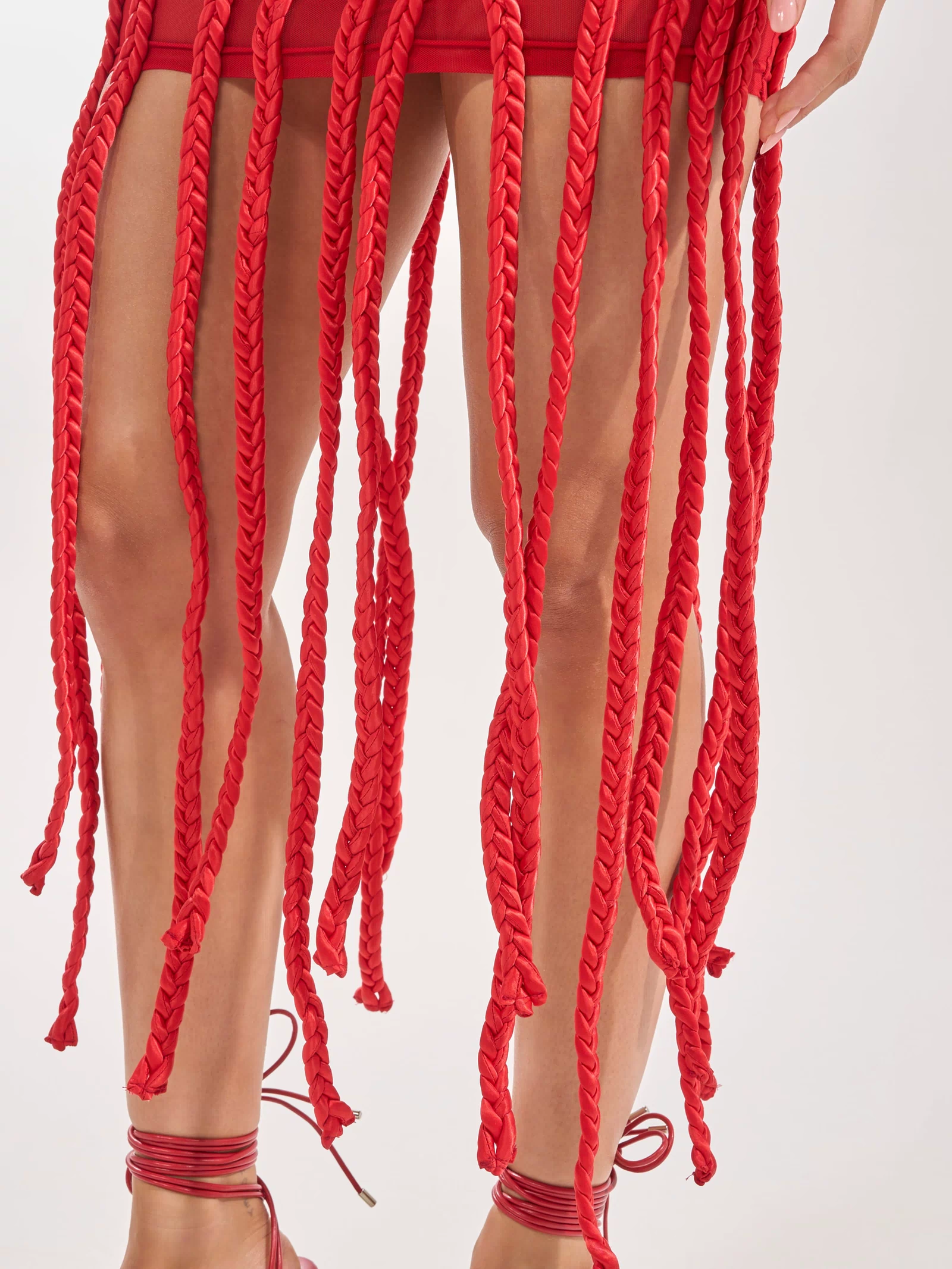 A person wears the STUCK ON YOU BRAIDED MINI DRESS IN RED, featuring long, thick braided red tassels, paired with red strappy sandals against a plain light-colored background.