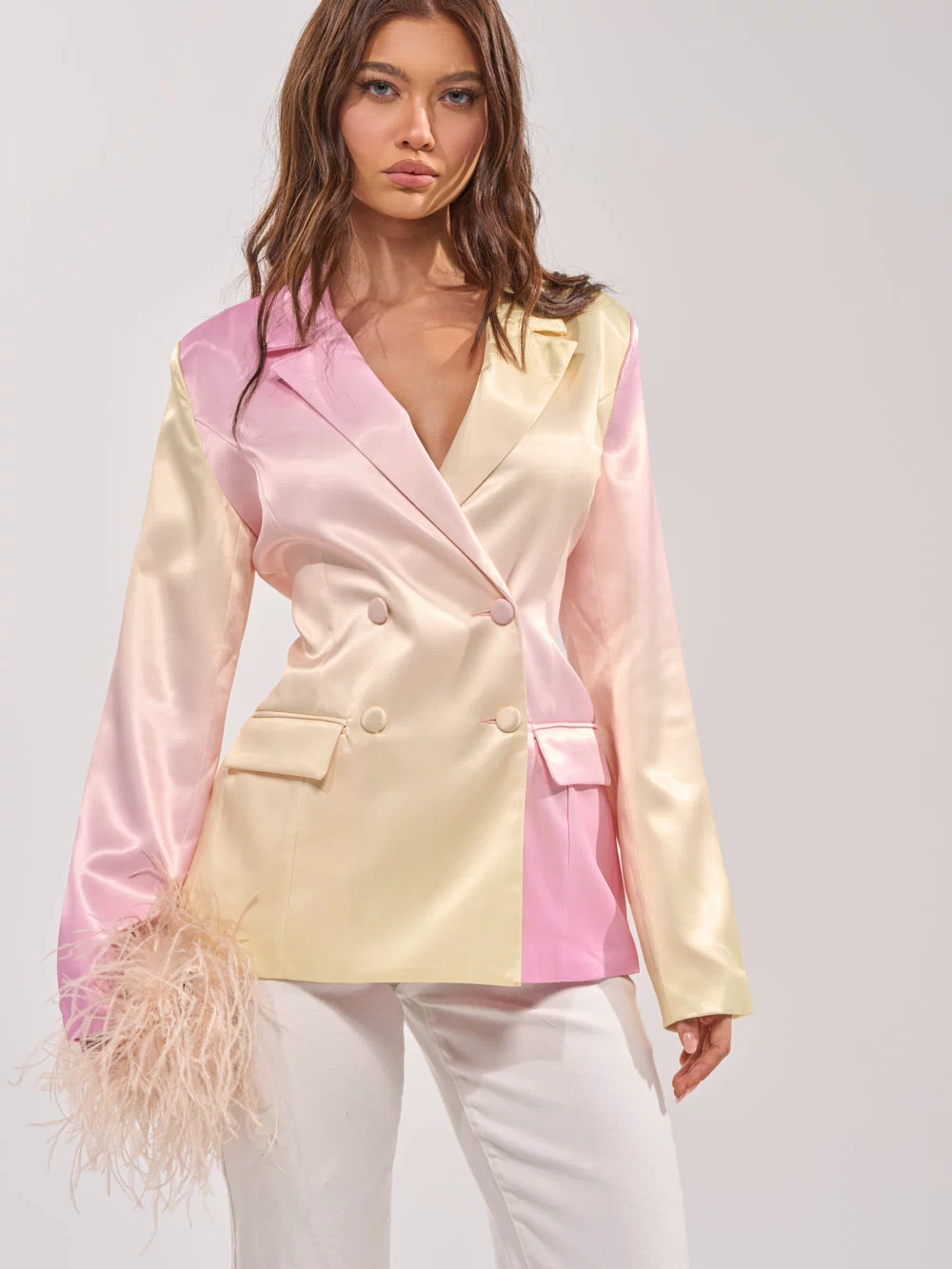 A woman models the SWEET TALK SATIN BLAZER in pastel pink and yellow blocks, paired with white pants and holding a beige feather accessory, standing against a plain light background.