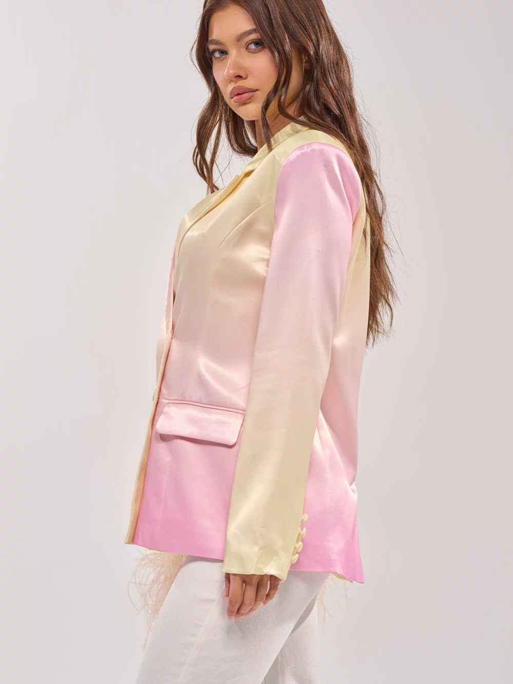 A woman with long brown hair models the SWEET TALK SATIN BLAZER in shiny pastel pink and yellow, paired with white pants and shown in side profile against a plain light gray background.