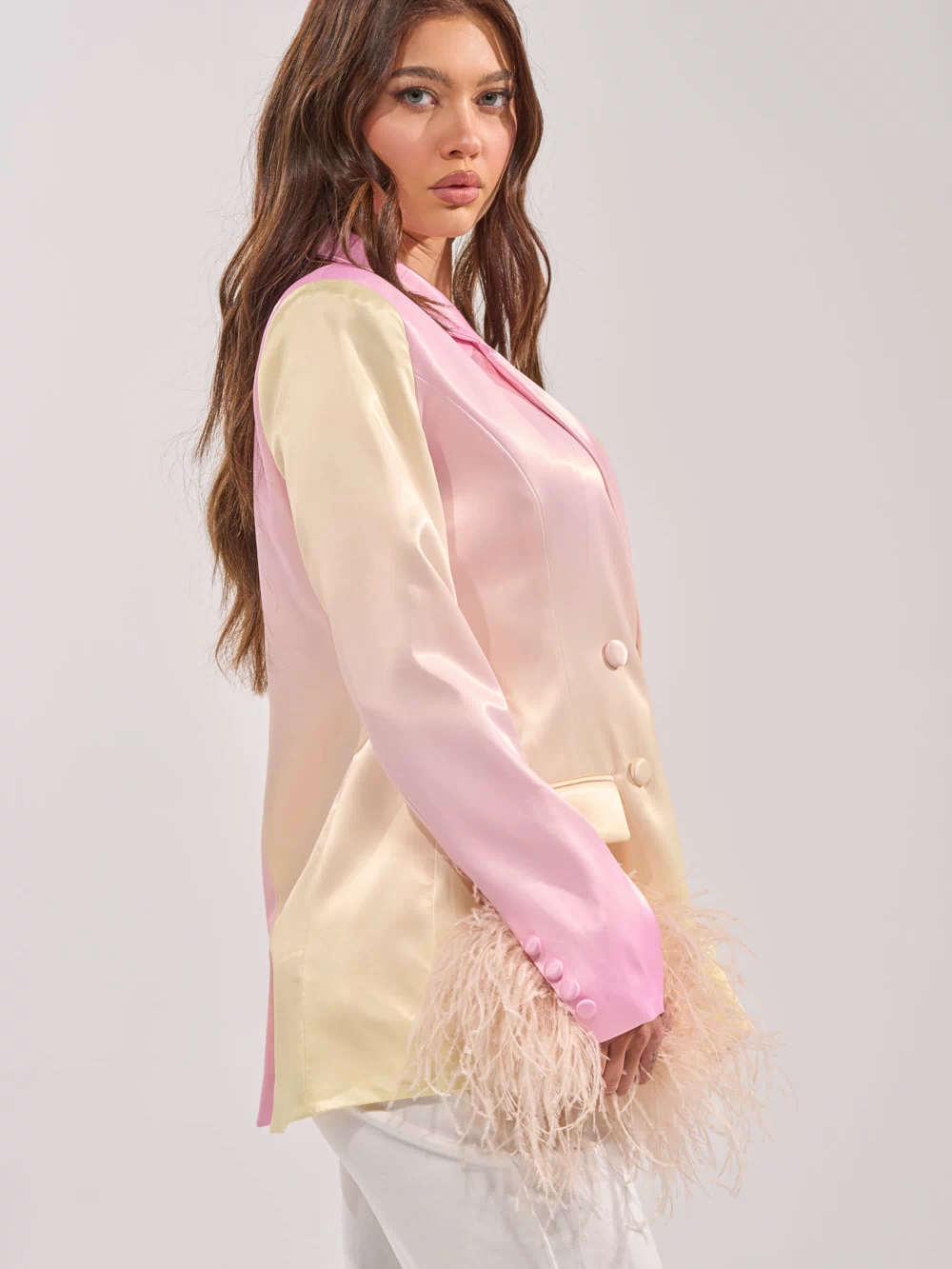 A woman with long wavy brown hair wears the SWEET TALK SATIN BLAZER with feather-detailed sleeves, paired with white pants, standing against a plain light background.