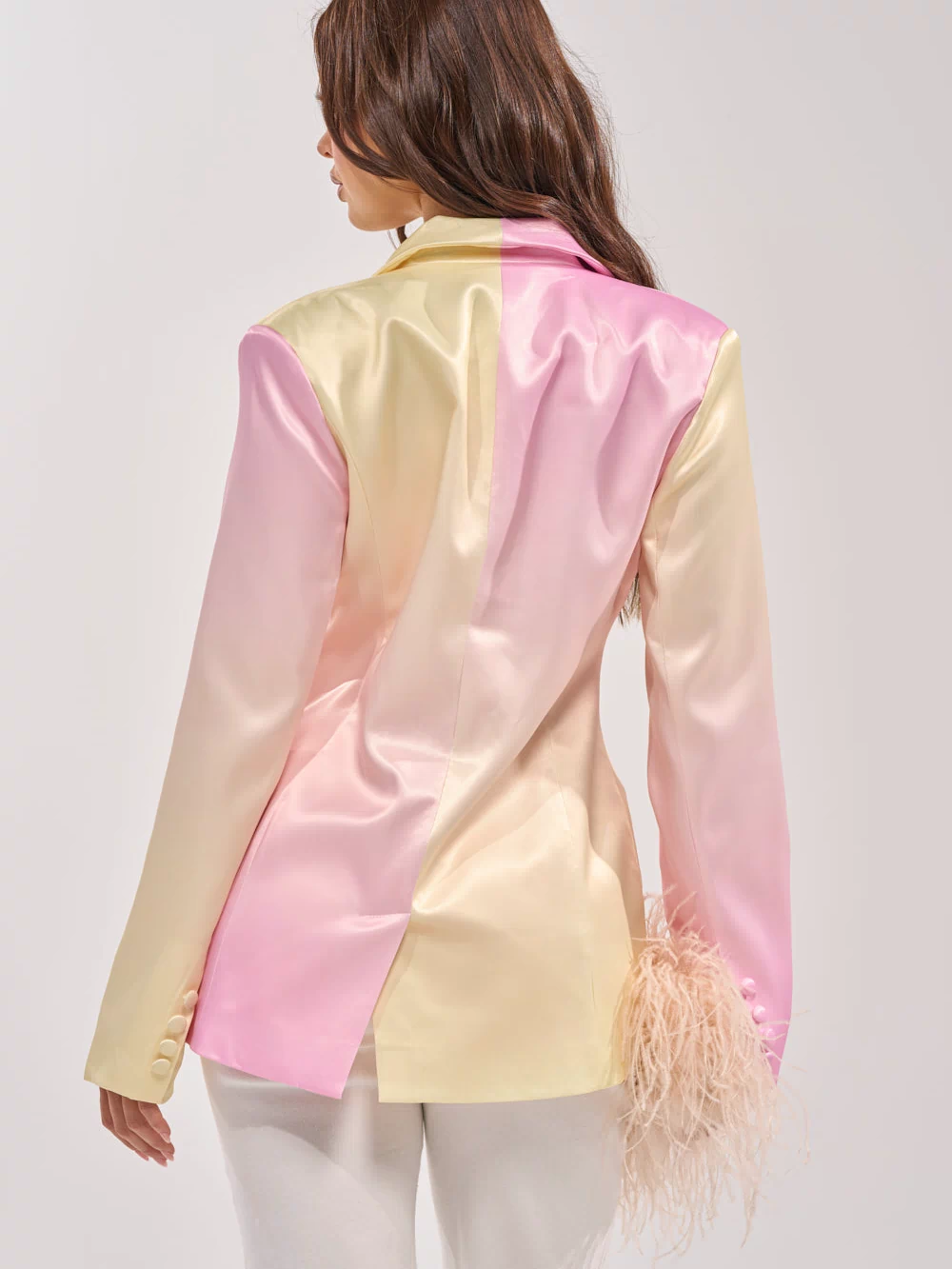 A woman with long brown hair models the SWEET TALK SATIN BLAZER in yellow and blush pink tones, seen from the back. The tailored blazer features a feather detail on one sleeve and is paired with crisp white pants.
