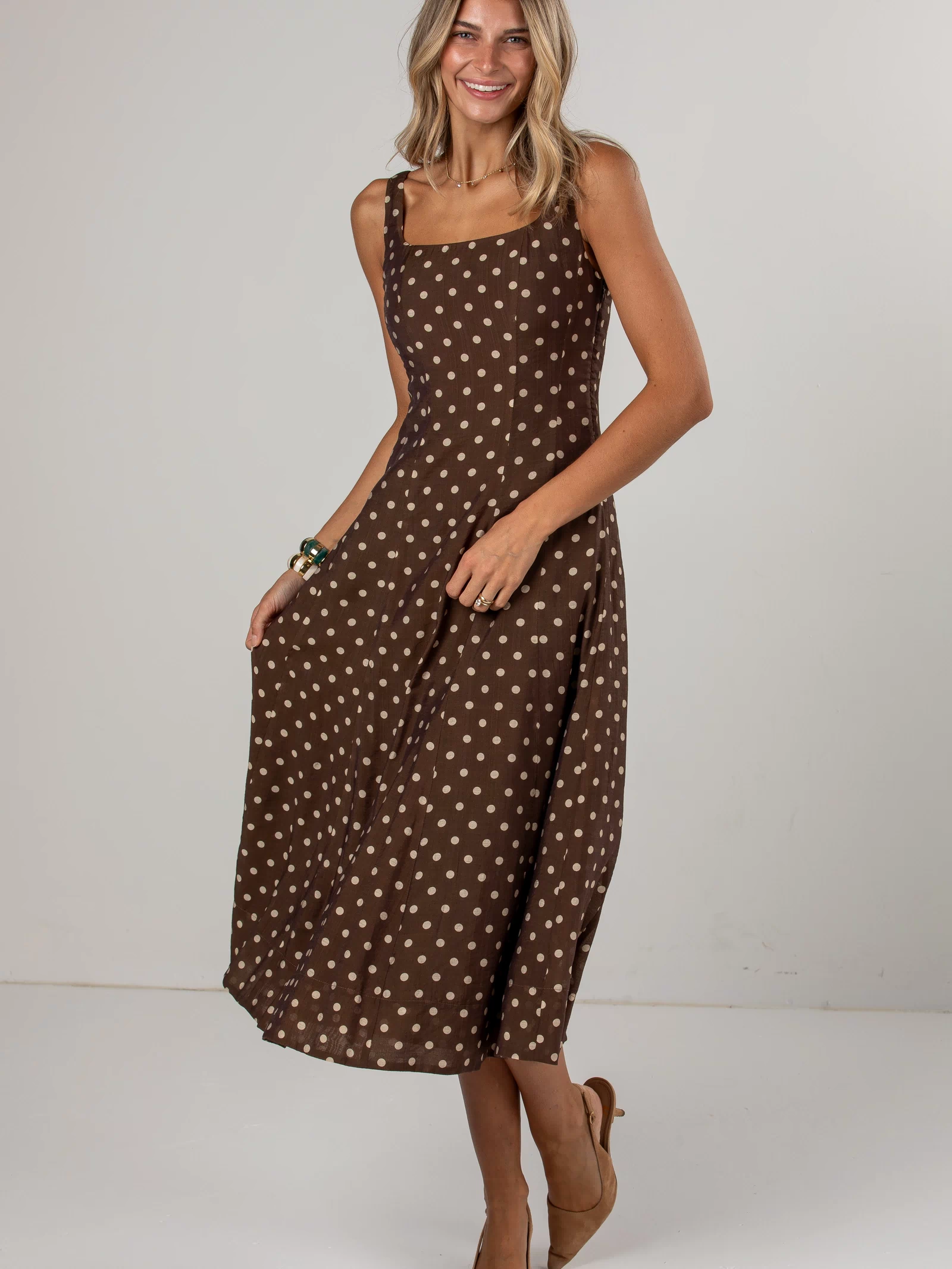 A woman modeling a brown midi dress with a white polka dot pattern, photographed against a neutral light gray studio background.