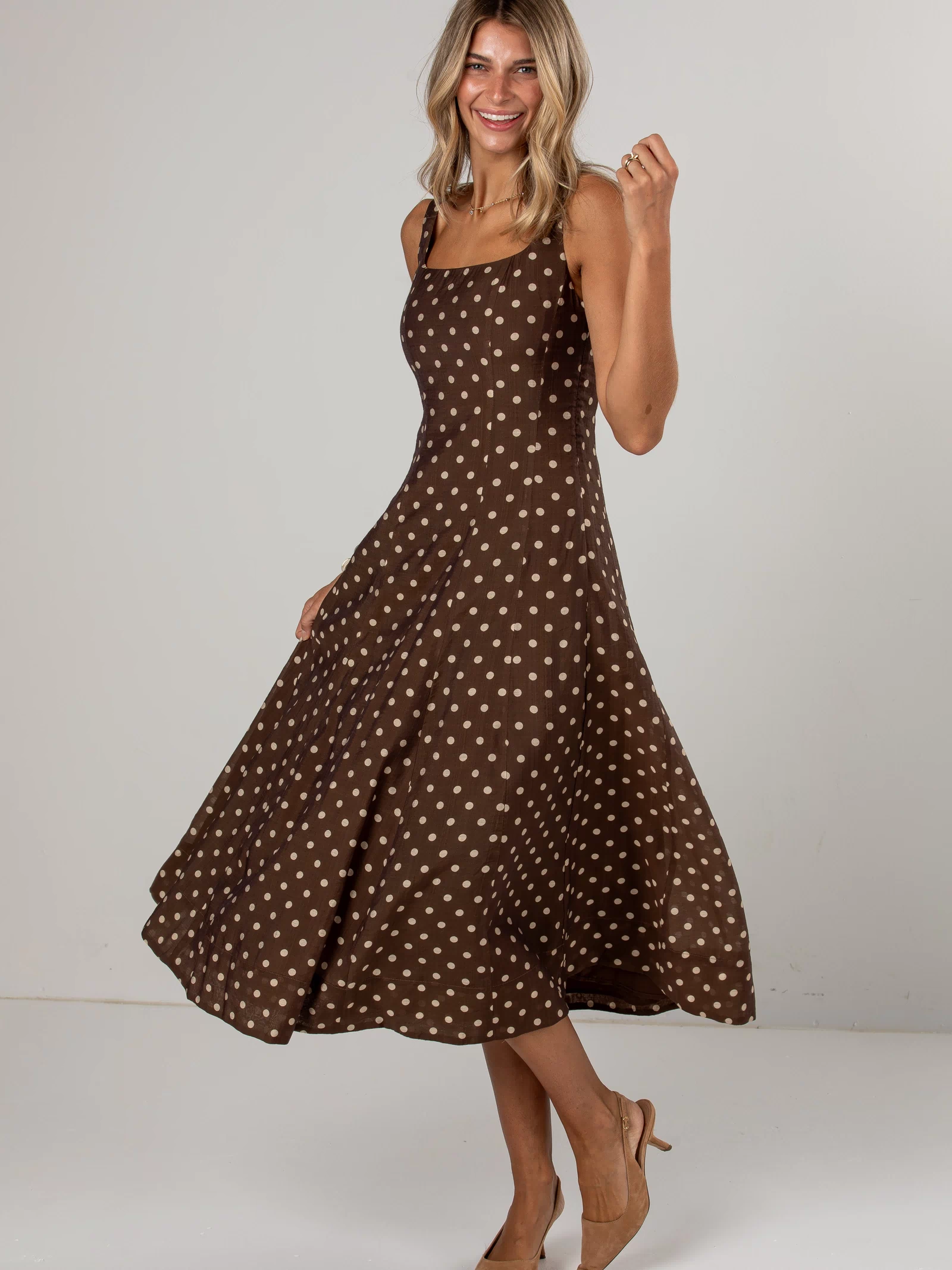 A woman modeling a brown midi dress with a beige polka dot pattern, standing in a studio setting against a light gray background.