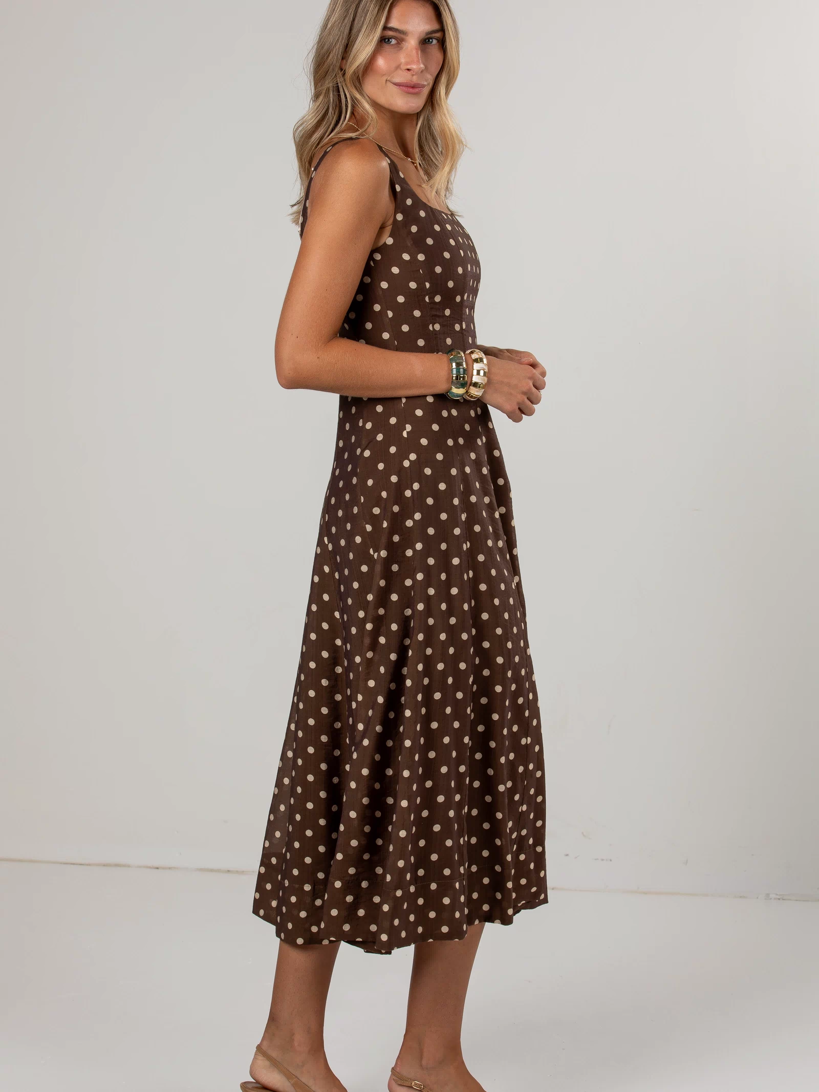 A woman modeling a sleeveless brown midi dress with a white polka dot pattern, paired with beige slingback heels, against a plain light gray studio background.