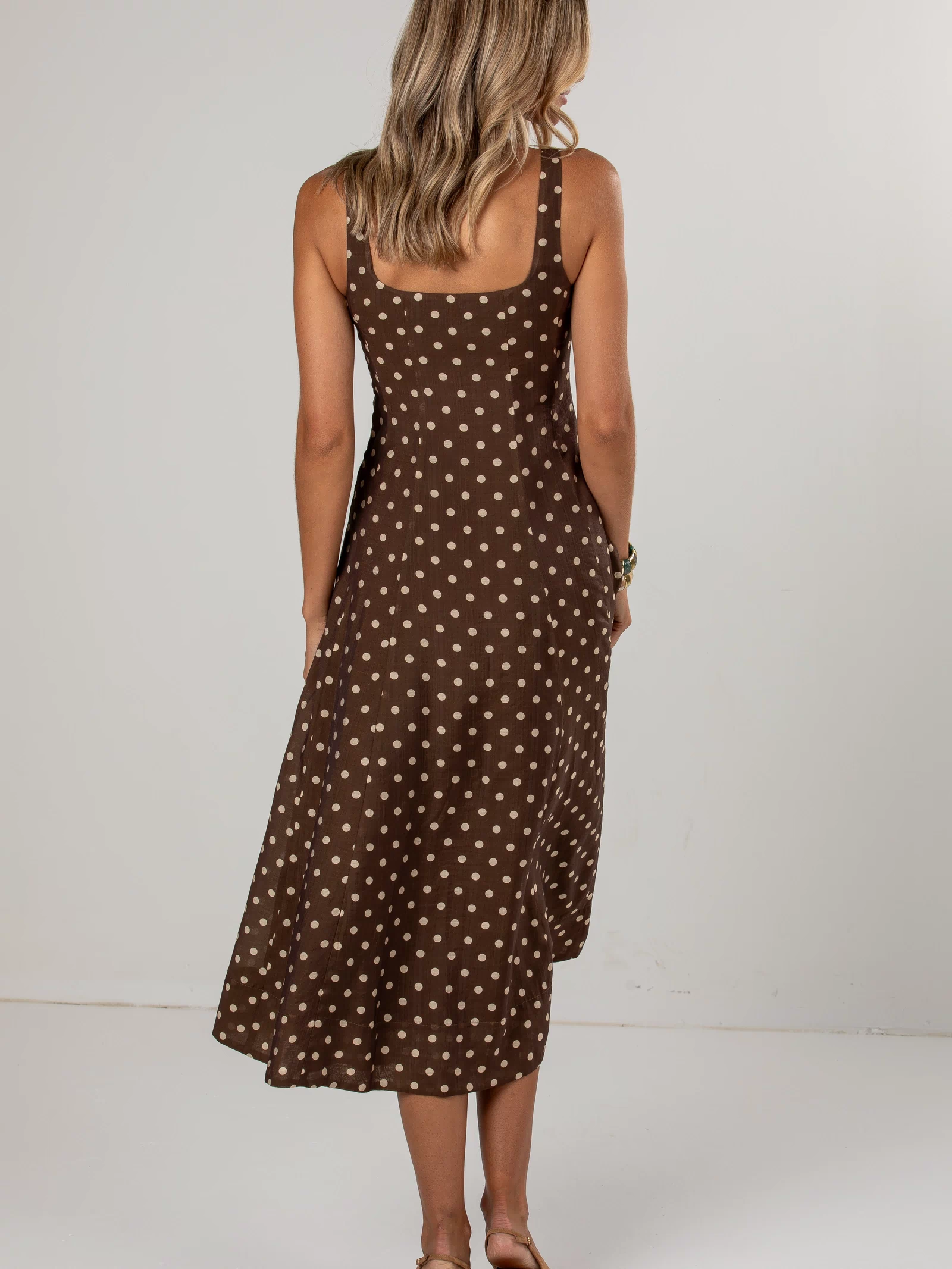 A woman viewed from behind wearing a brown midi dress with a white polka dot pattern, paired with tan heeled sandals, set against a plain light gray studio background.