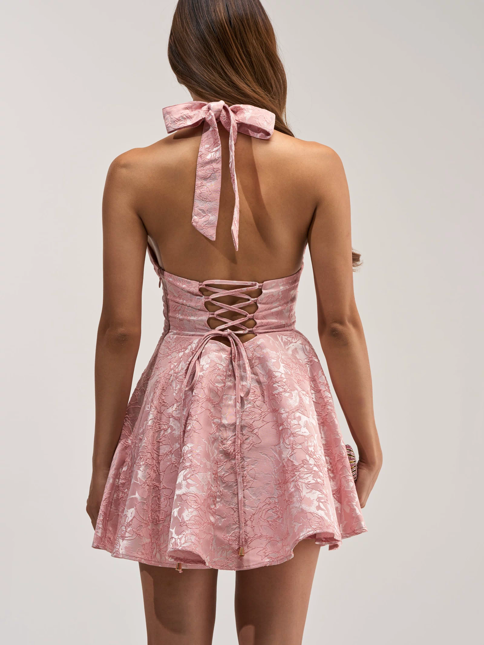 A woman is seen from behind wearing the TALK IS CHEAP FLORAL MINI DRESS—a shiny pink, floral-patterned halter mini dress featuring a lace-up back and flared skirt.