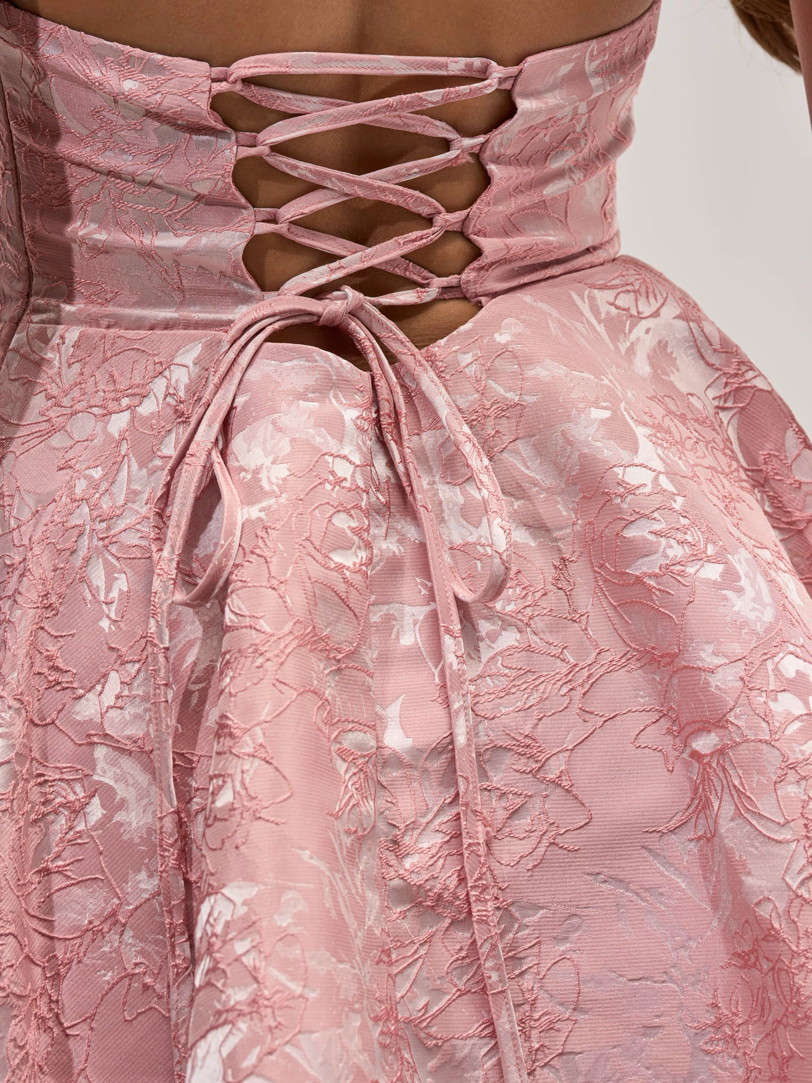 Close-up of the TALK IS CHEAP FLORAL MINI DRESS, showcasing pink floral embroidery and brocade detailing, with a lace-up corset-style back and a bow tied at the waist, worn by a person with bare shoulders.