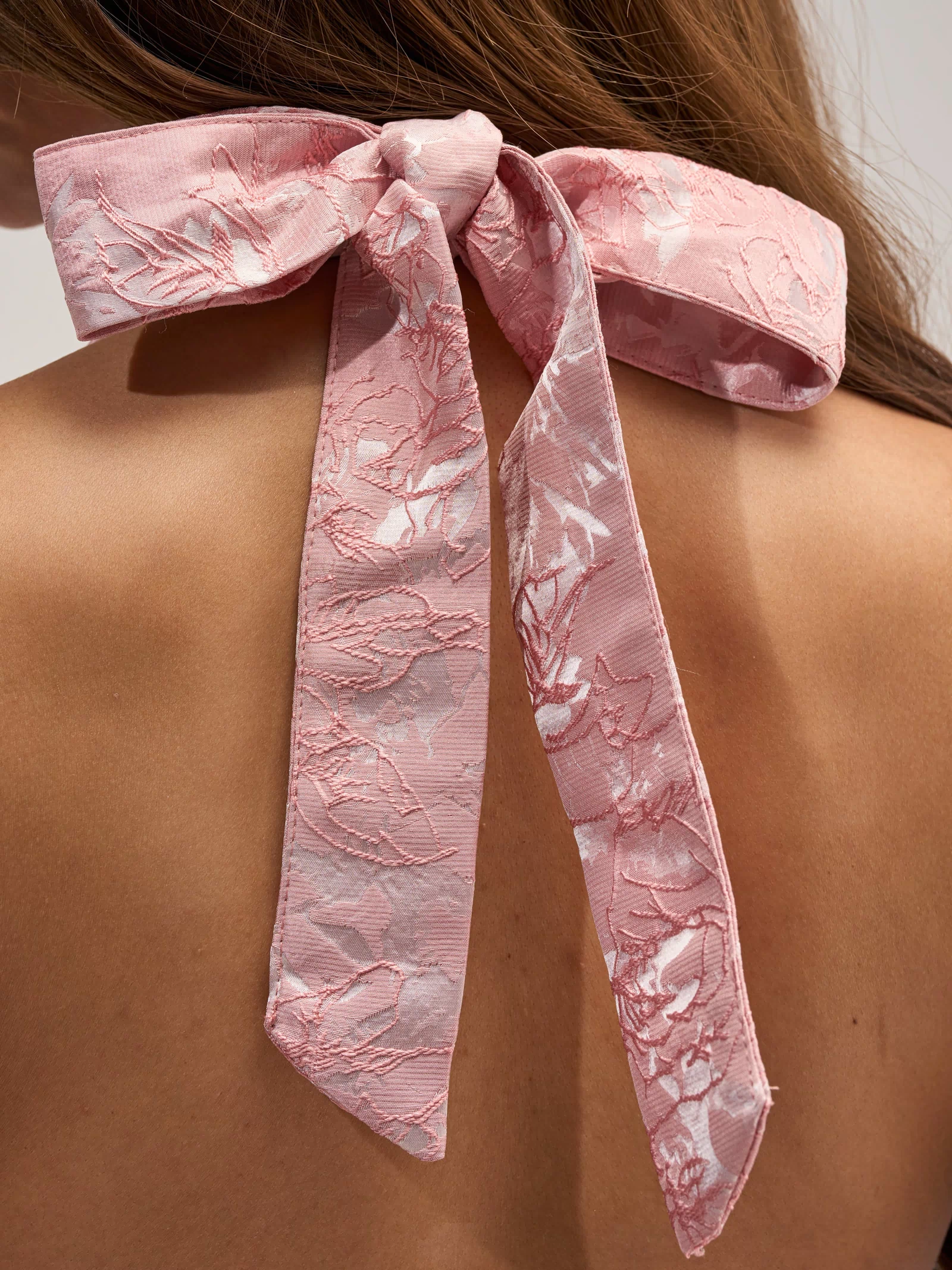 Close-up of a person’s back with long hair, showing a large pink textured bow tied at the neckline of the TALK IS CHEAP FLORAL MINI DRESS.
