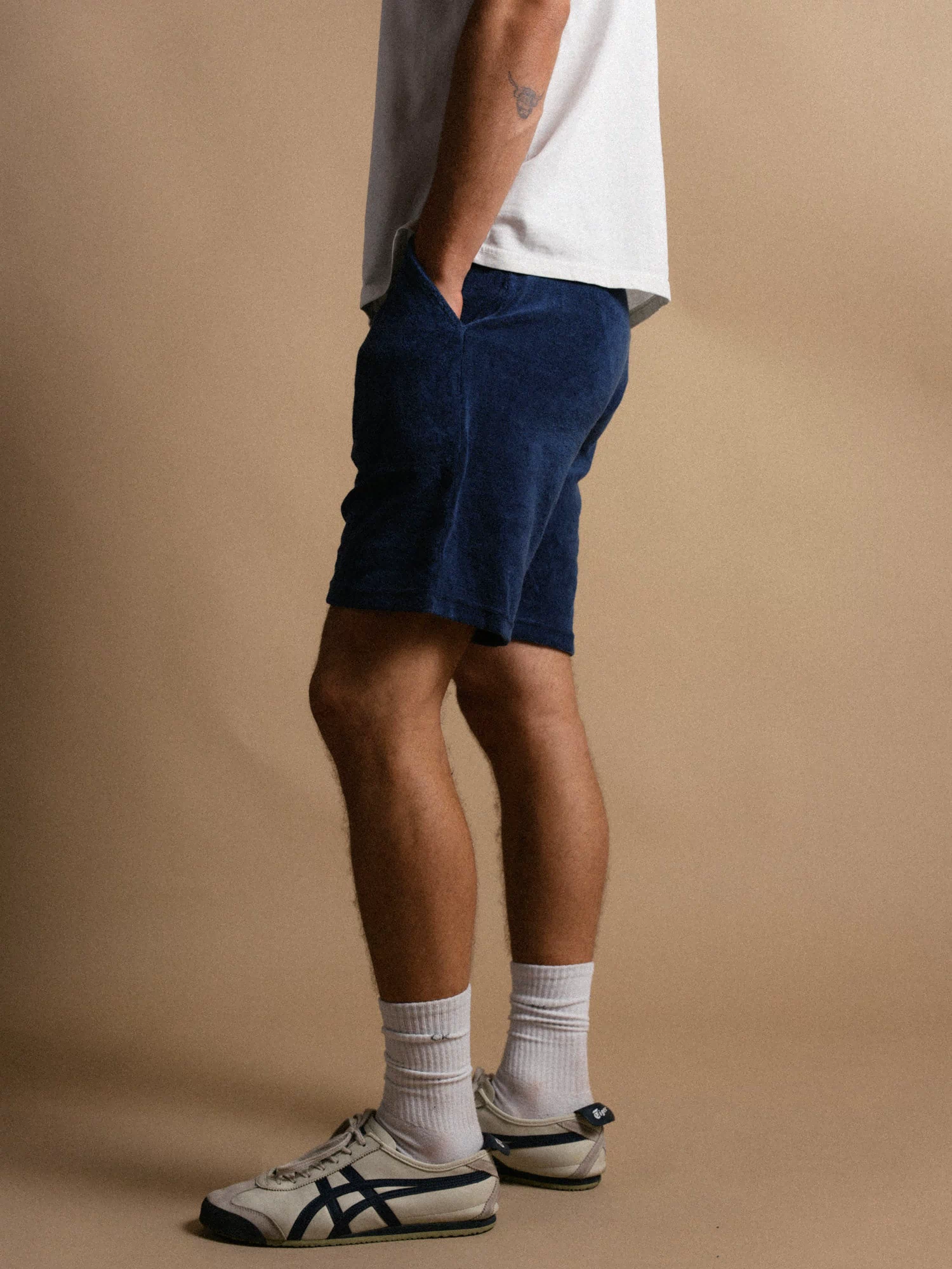 Man in white shirt, blue shorts, and sneakers stands against a beige background, hands in pockets, showcasing casual style.