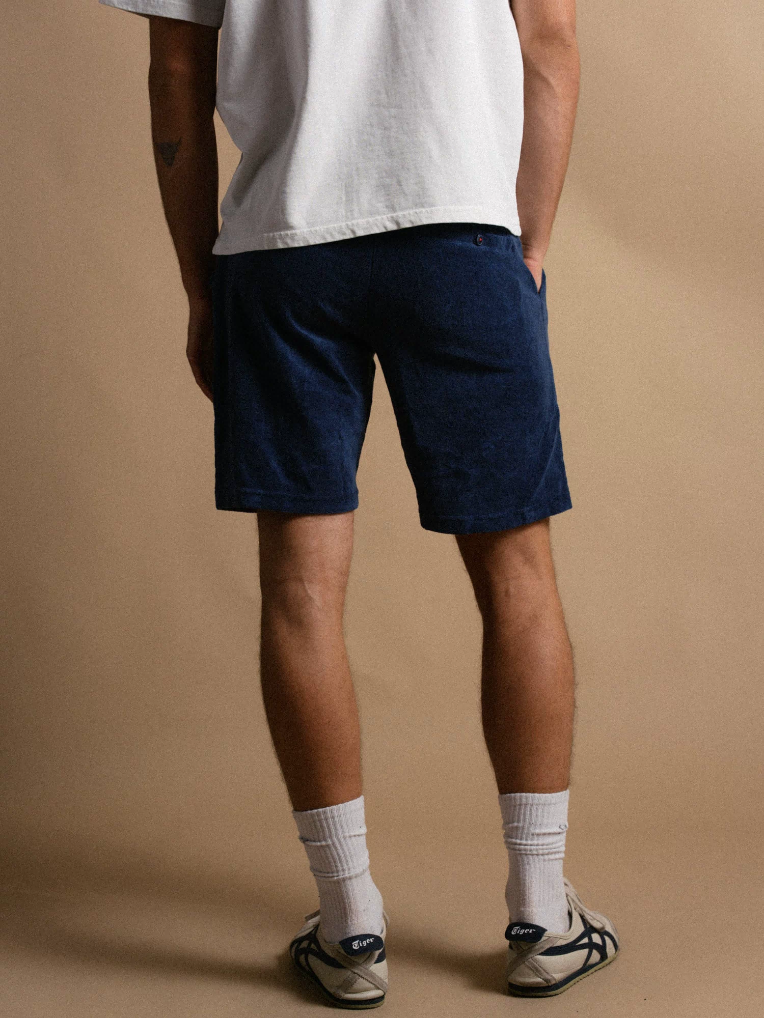 Man in white shirt and blue shorts stands against a beige background, wearing white socks and sneakers.