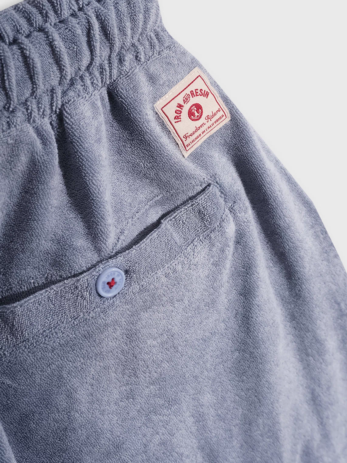 Blue terry cloth shorts with a buttoned back pocket and red "Iron and Resin" label, showcasing casual style and comfort.