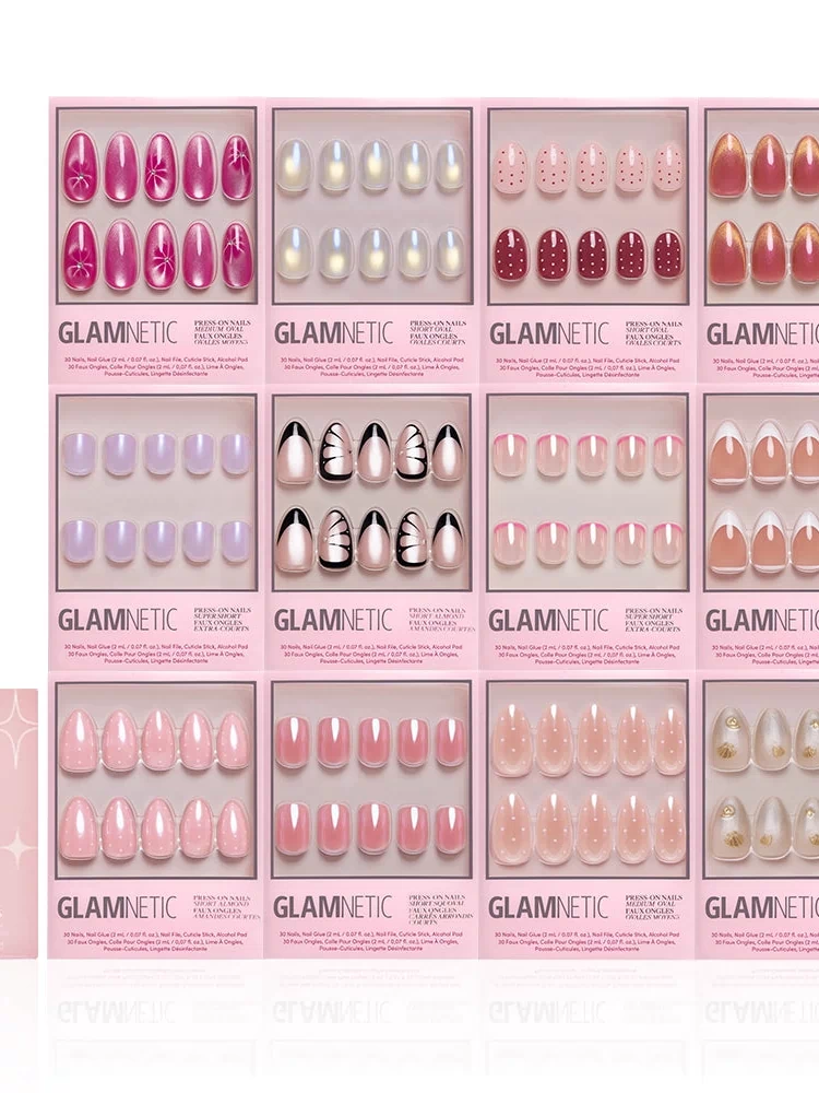 Twelve sets of glamnetic’s The Spring Edit Collection press-on nails, in pink boxes with various shapes, colors, and shimmery designs, are arranged in a grid beside a pink nail application kit on the lower left.