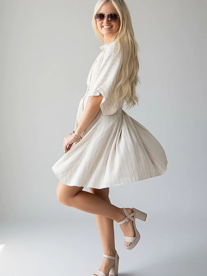 A woman modeling a short-sleeve cream linen dress with a flared skirt, paired with beige platform sandals and sunglasses, posing against a light gray studio background.
