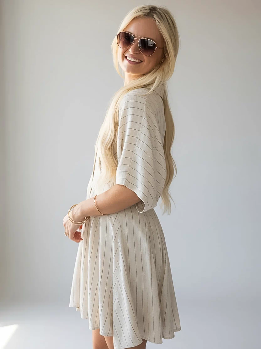 A blonde woman wearing a cream-colored pleated short-sleeve dress and sunglasses, posing in a studio setting against a light gray background.